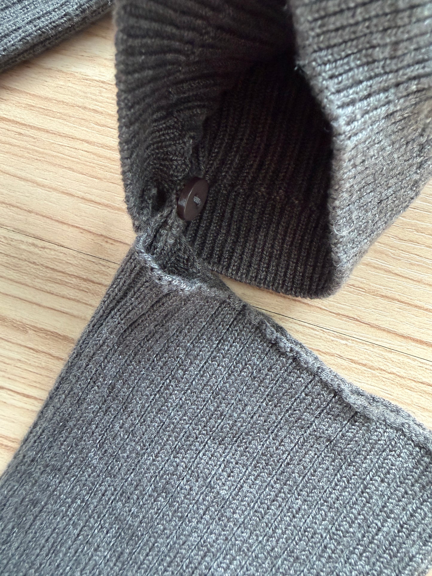 Close-up of a dark gray ribbed knit garment laid on a light wood surface, showing the sleeve cuff, button, and fabric texture.