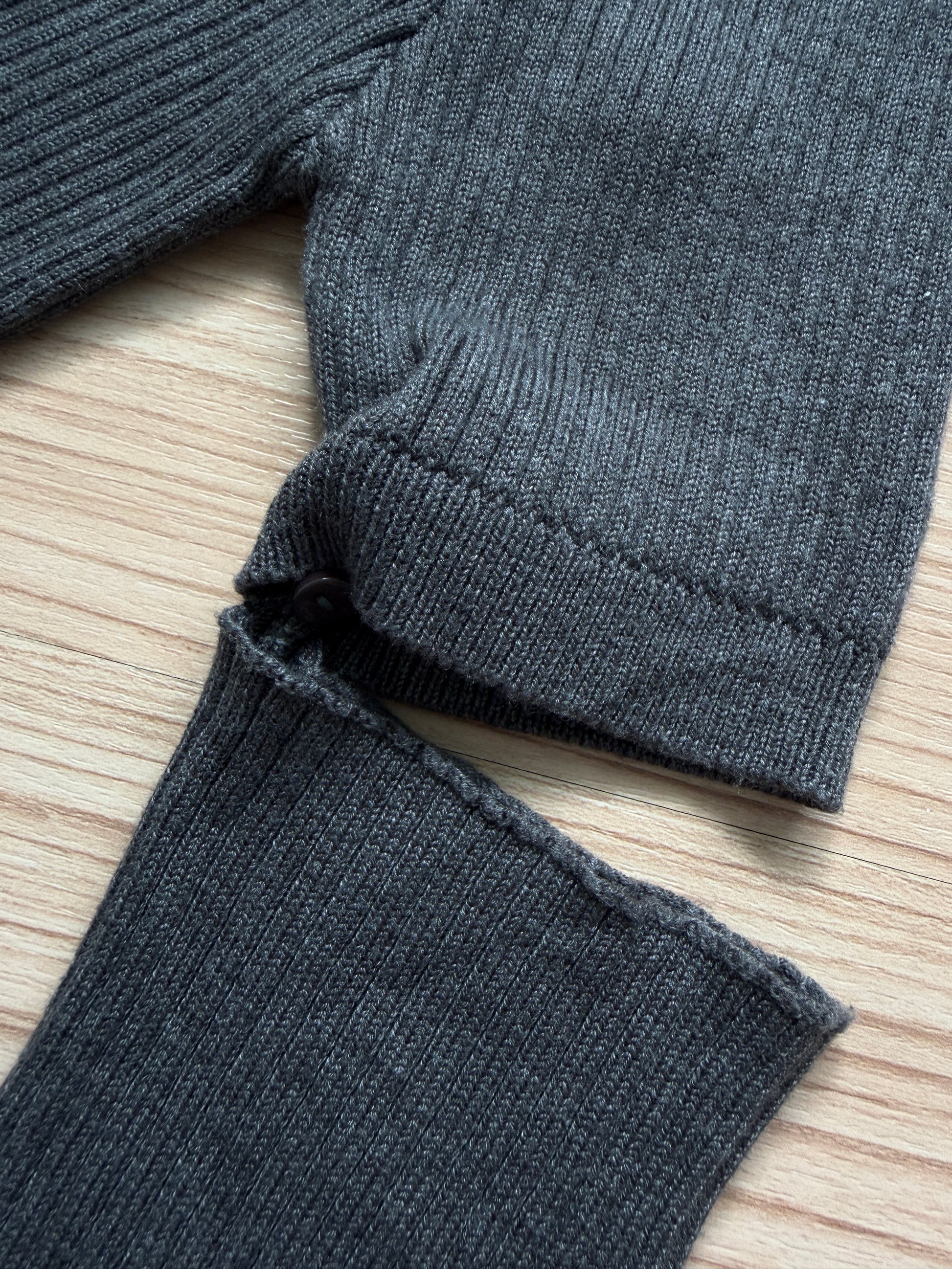 Close-up of a dark gray ribbed knit garment laid flat on a light wood surface, showing the sleeve cuff and lower hem detail.