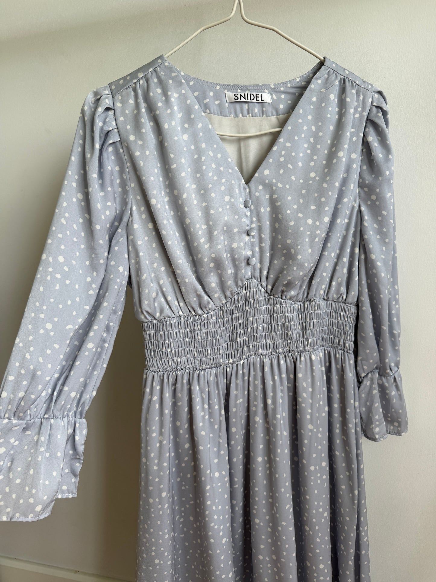 A light gray long-sleeve dress with a white polka dot pattern, featuring a smocked waist and ruffled cuffs, hanging on a white hanger against a plain beige wall.