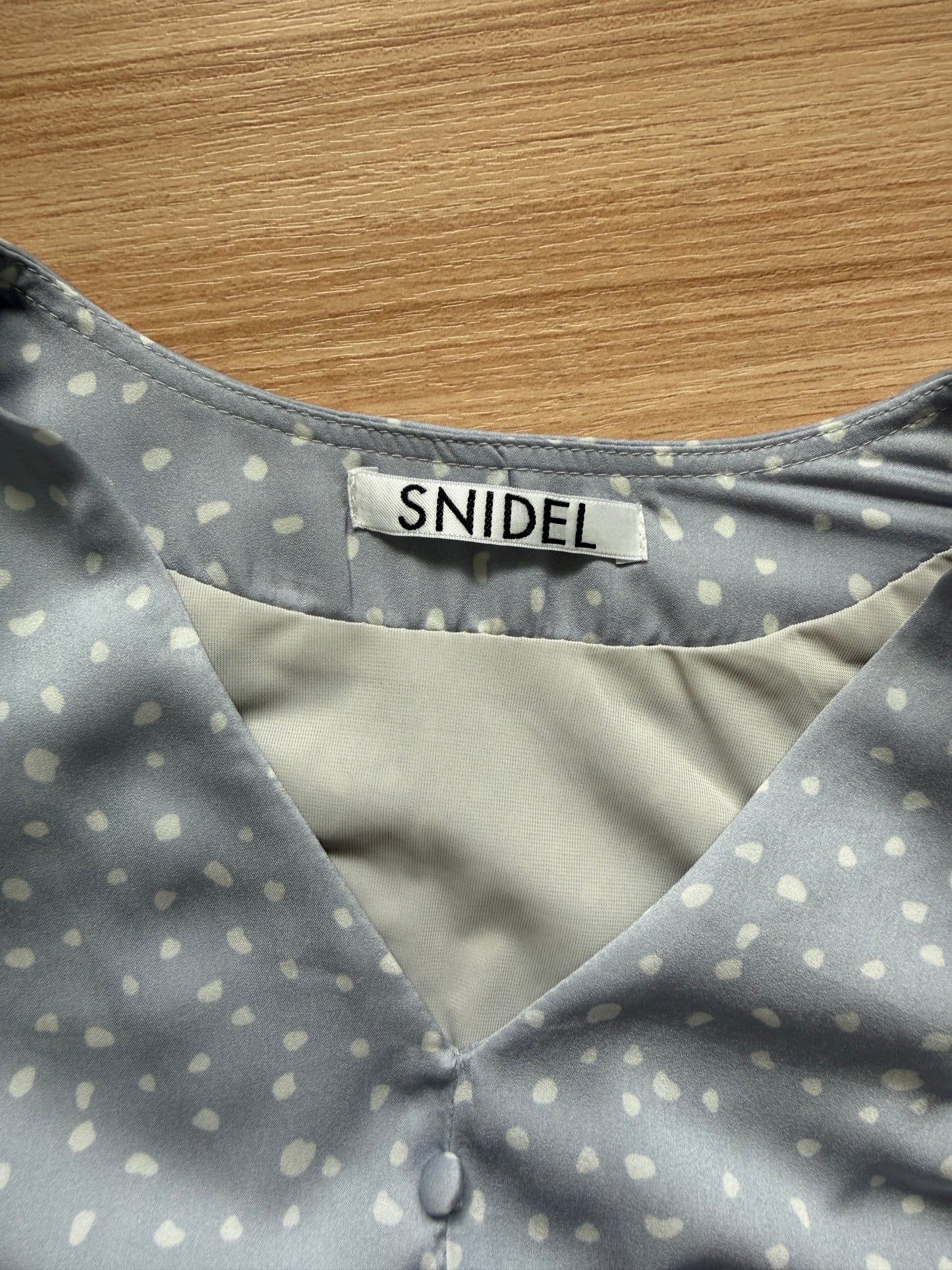 Close-up of a Snidel blouse with a gray and white polka dot pattern, featuring a V-neckline and a visible brand label.
