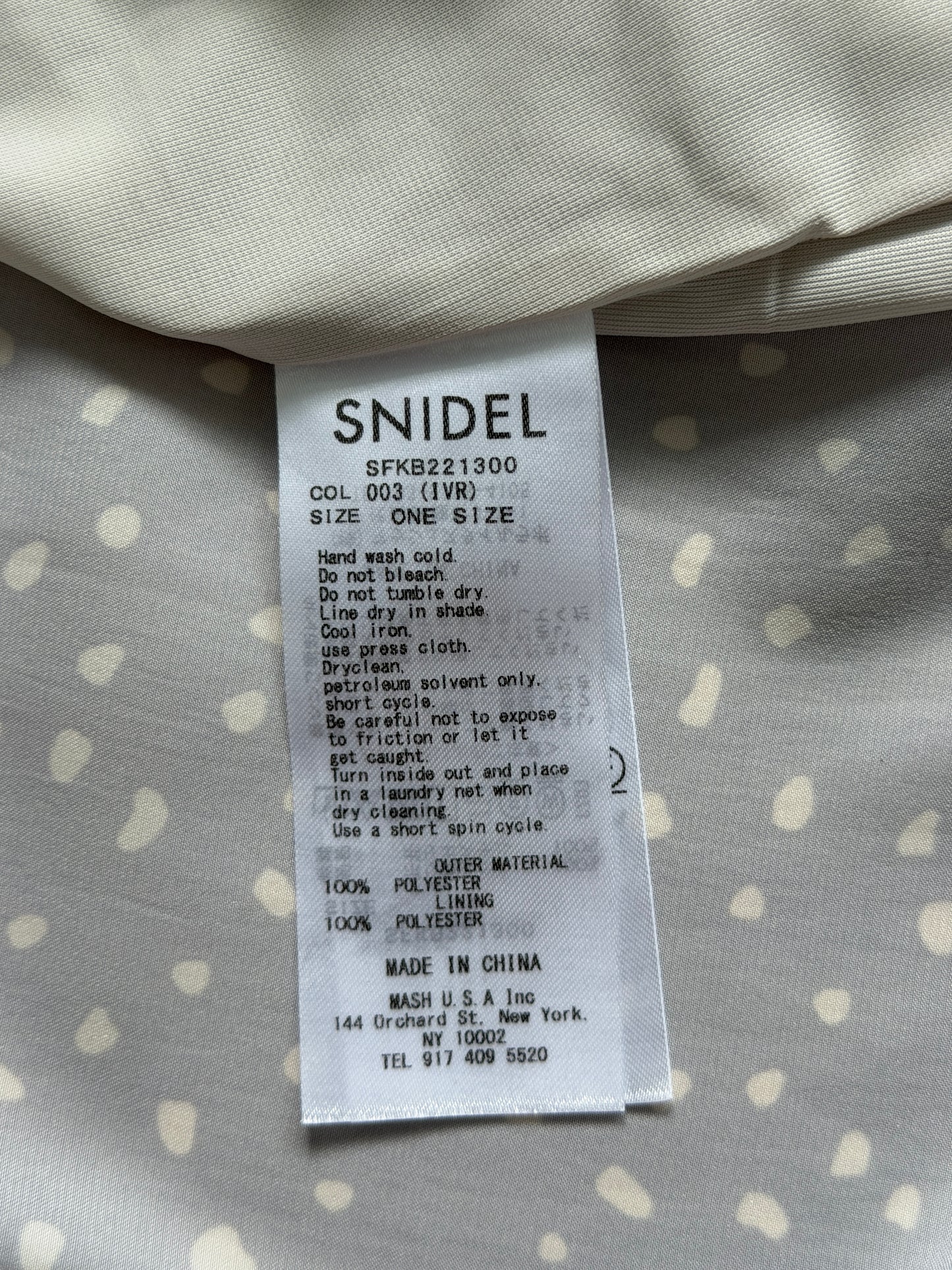 A close-up of a white fabric care label reading 'SNIDEL' with product details, placed on a light gray fabric with small white polka dots.