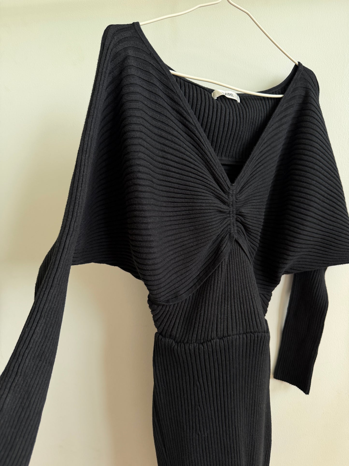 A black ribbed knit long-sleeve top with a twisted front detail, hanging on a white hanger against a plain beige wall.