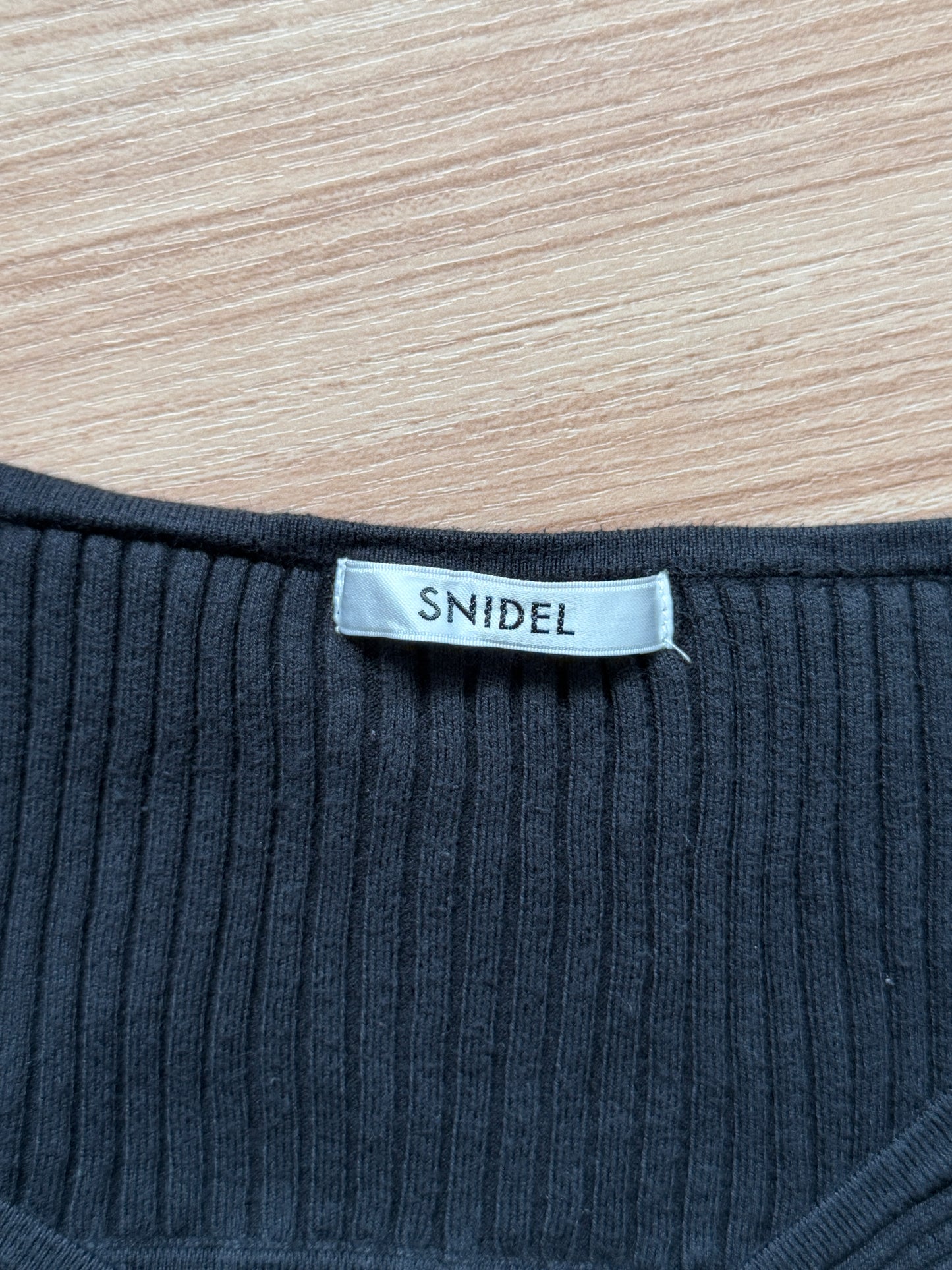 Close-up of a ribbed knit garment neck with a visible fabric label reading 'SNIDEL' on a light wood surface.