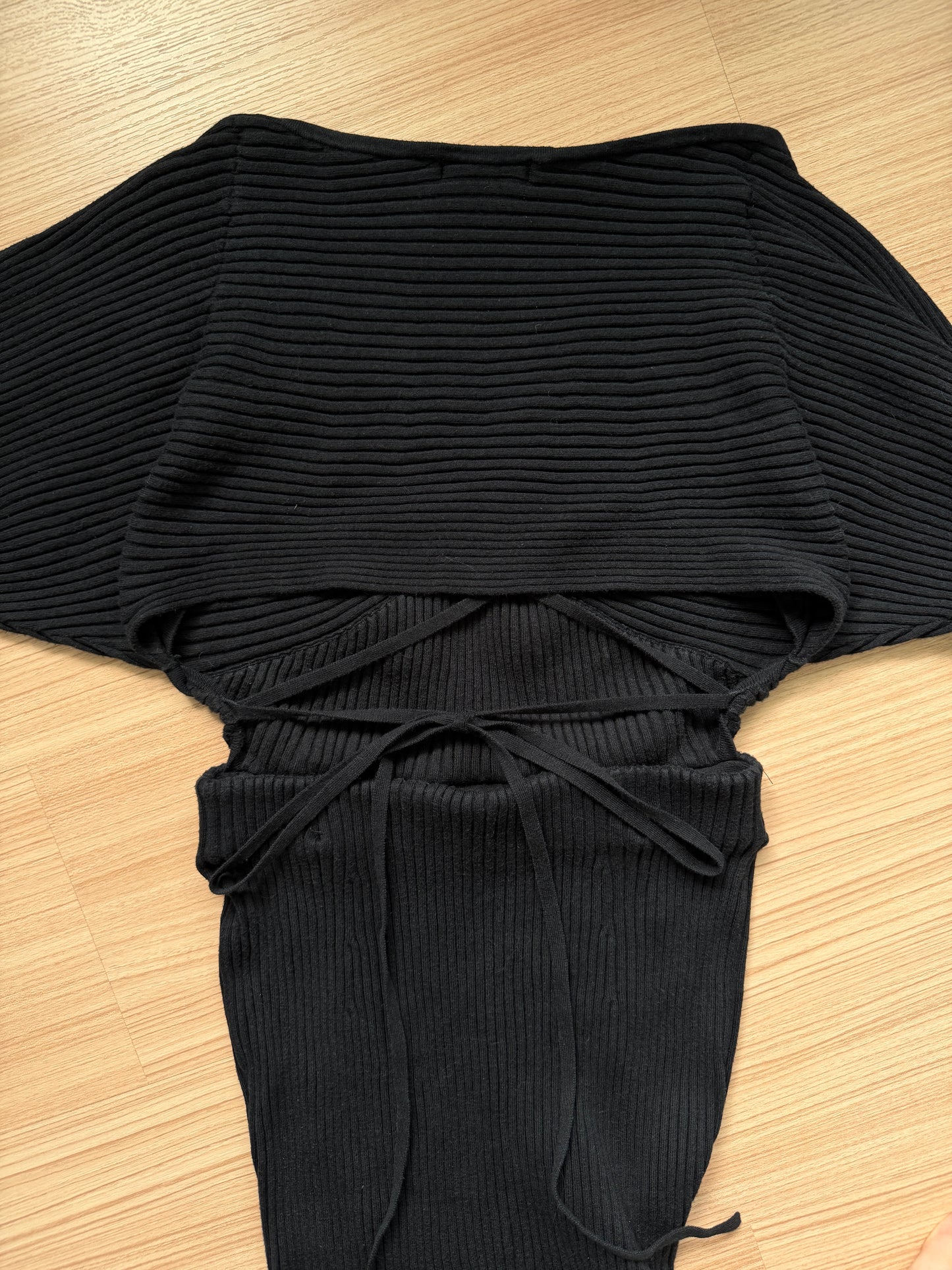 A black ribbed knit top with wide batwing sleeves, a high neckline, and a drawstring tie at the waist, laid flat on a light wood floor.