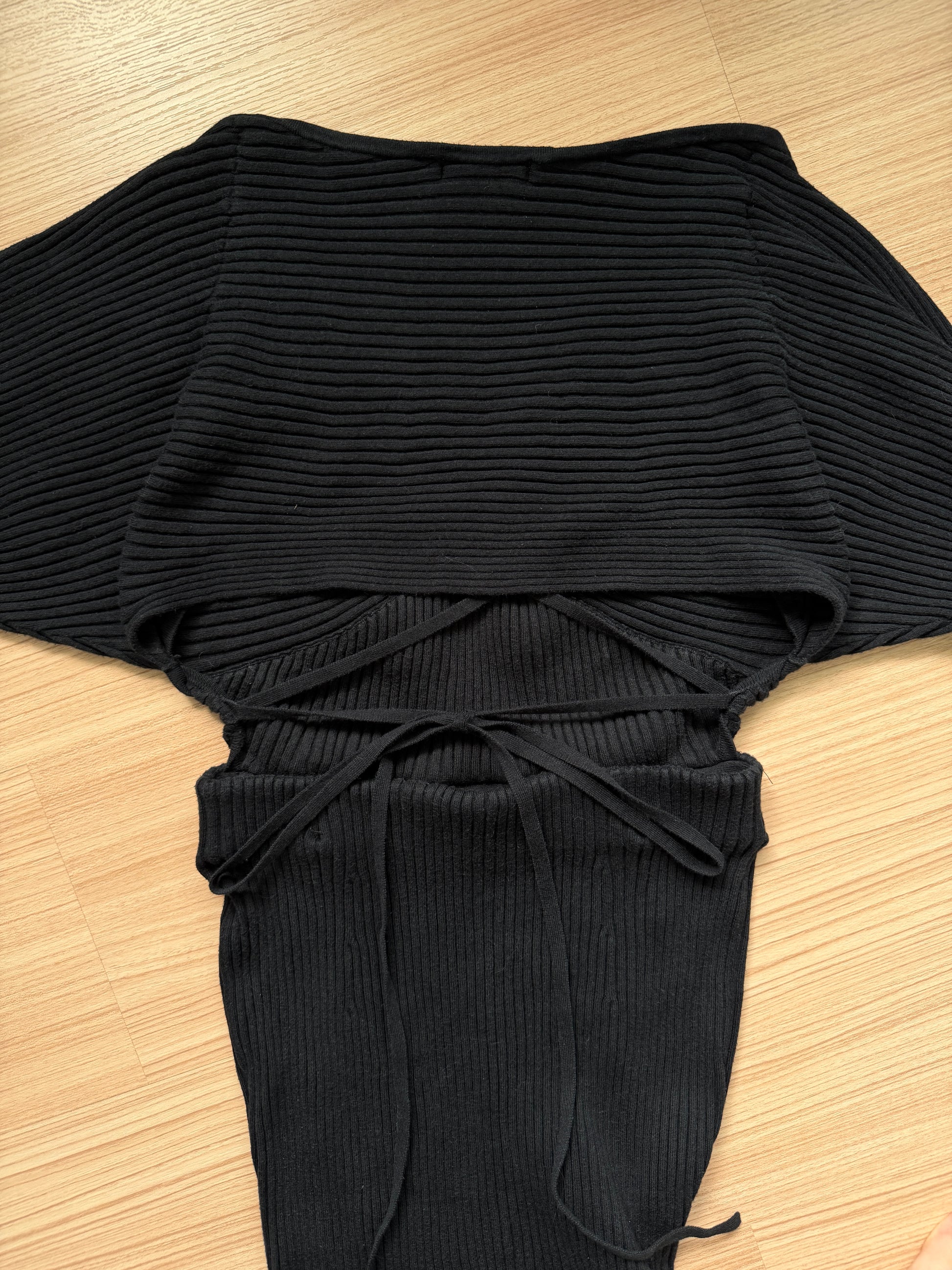 A black ribbed knit top with wide batwing sleeves, a high neckline, and a drawstring tie at the waist, laid flat on a light wood floor.