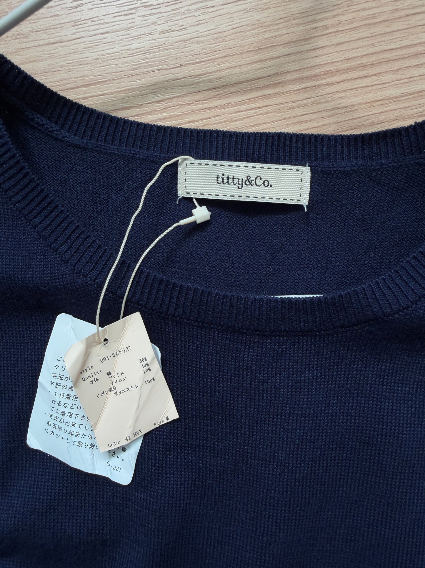 Close-up of a navy knit sweater with a visible neck label reading 'titty&Co.' and hanging product tags on a light wood surface.