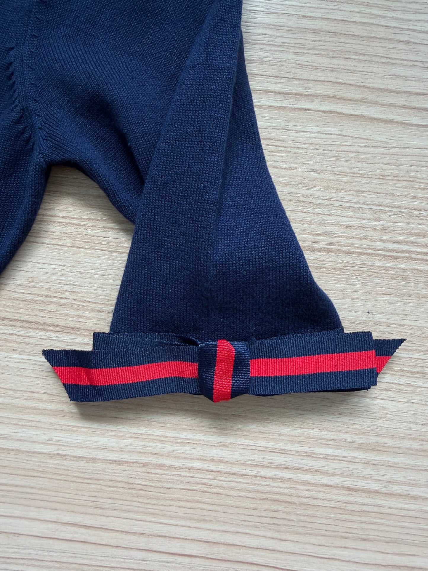 Close-up of a navy blue knit garment, likely a hat or scarf, folded on a light wood surface. The lower edge features a decorative bow made from a matching navy ribbon with a bright red stripe.