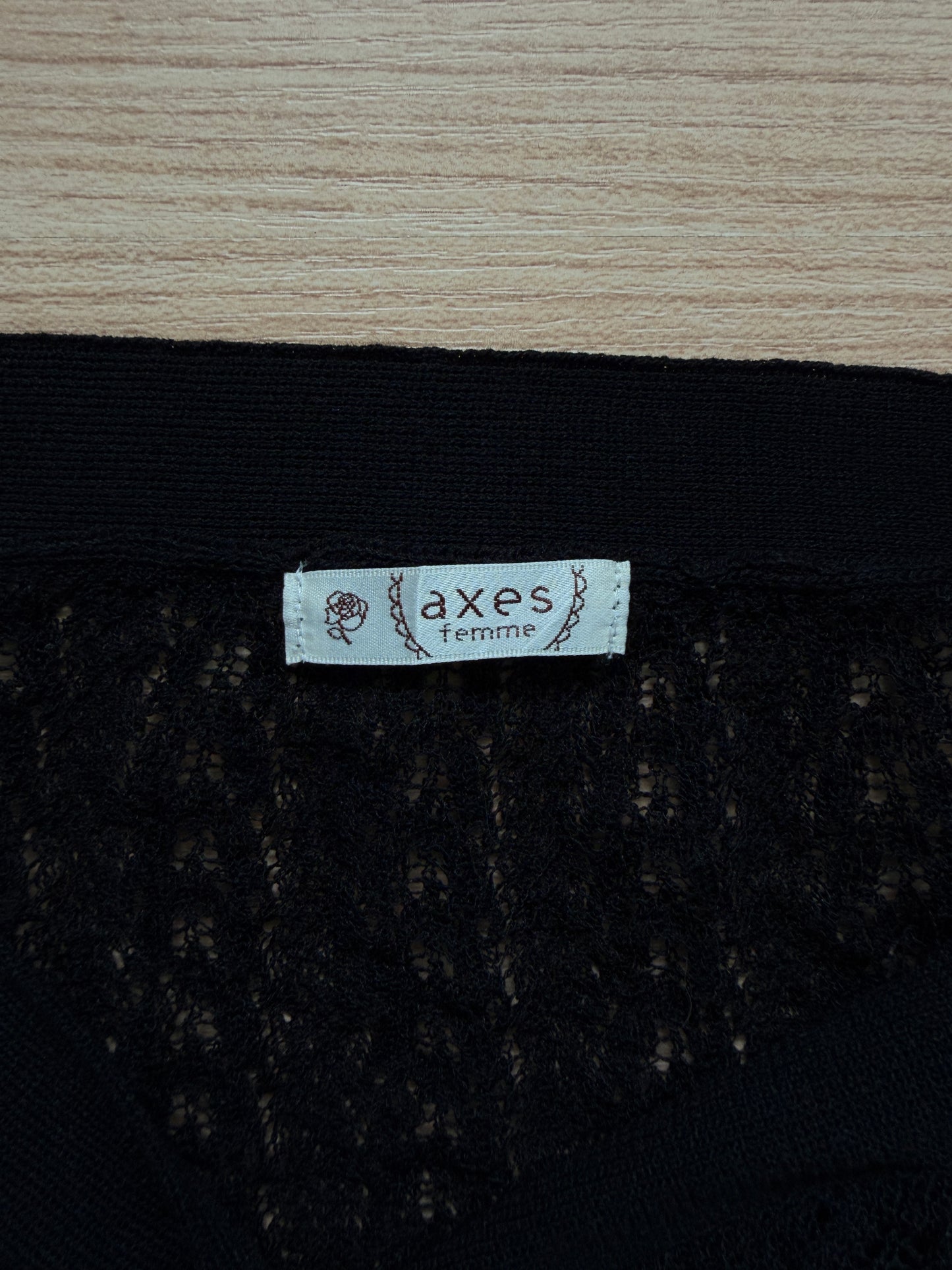 Close-up of a black lace garment with a white fabric label reading 'axes femme' on a light wood surface.