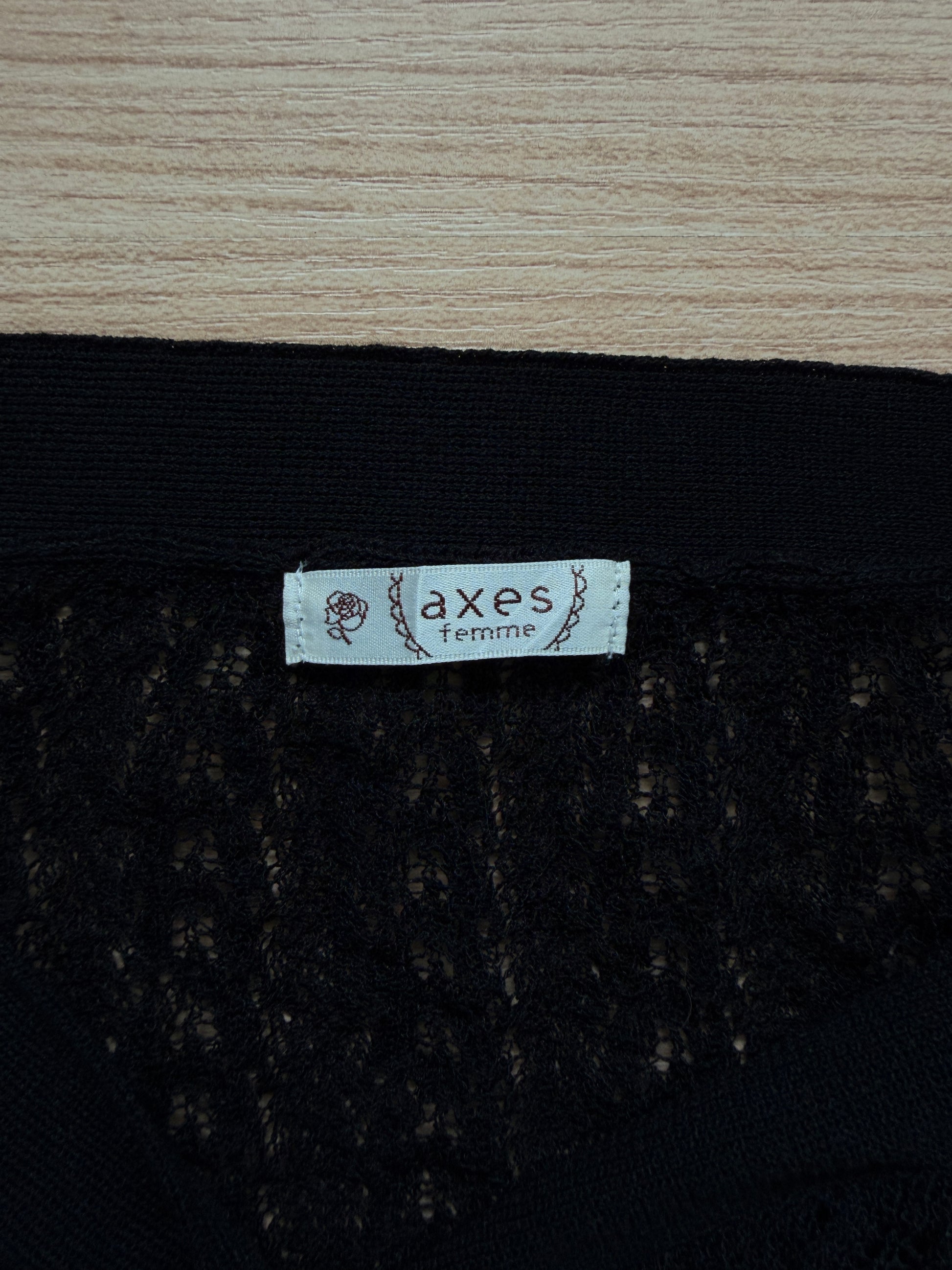 Close-up of a black lace garment with a white fabric label reading 'axes femme' on a light wood surface.