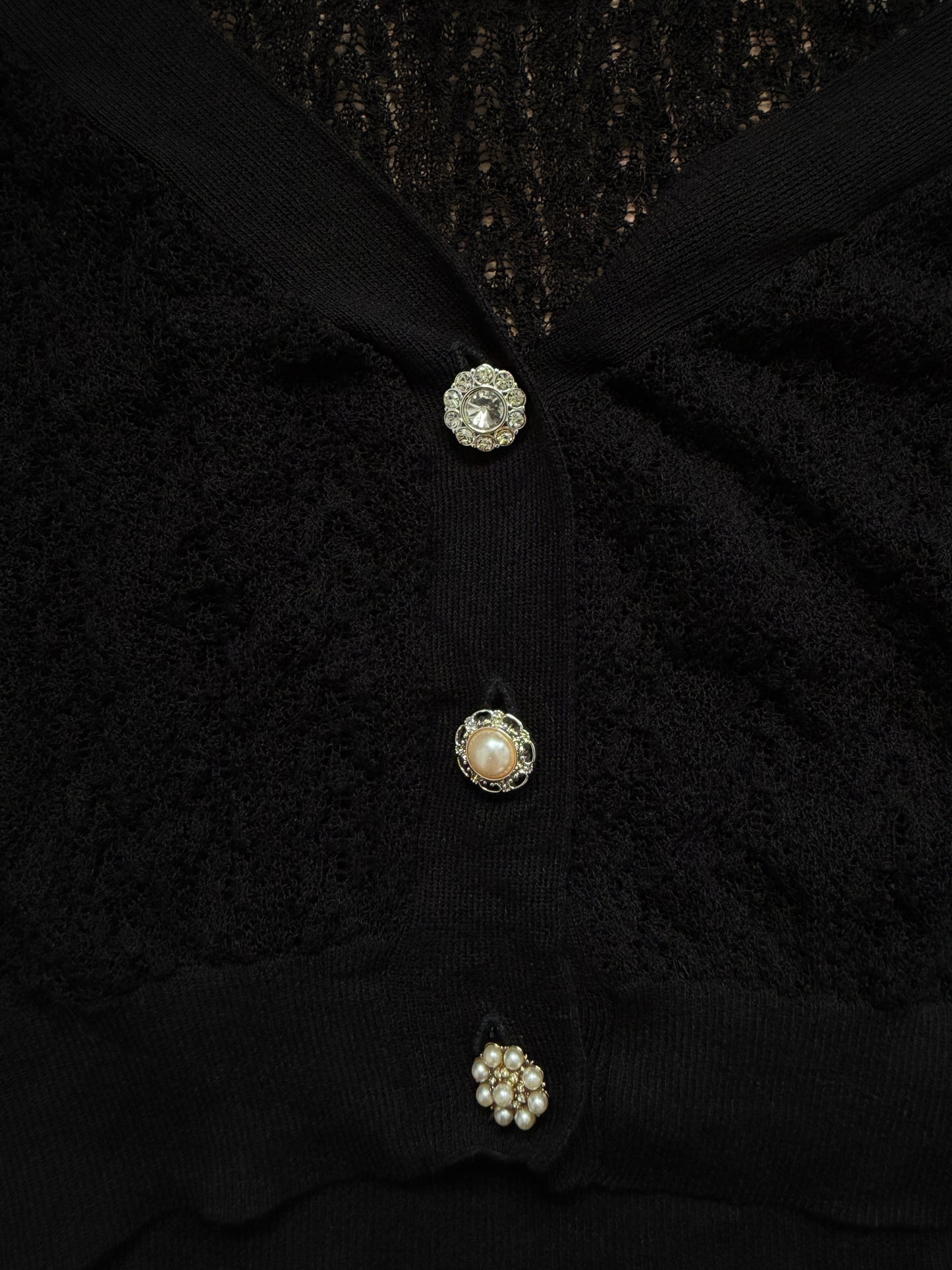 Close-up of a black textured knit cardigan with three decorative buttons featuring pearls and metallic detailing.