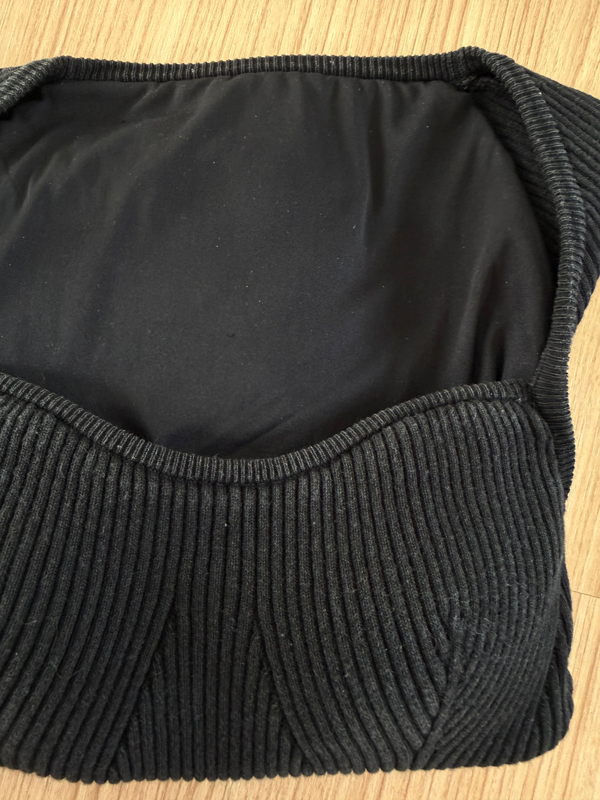 A close-up of a black ribbed knit bralette or bandeau-style top laid flat on a light wood surface, showing the underband and one cup.