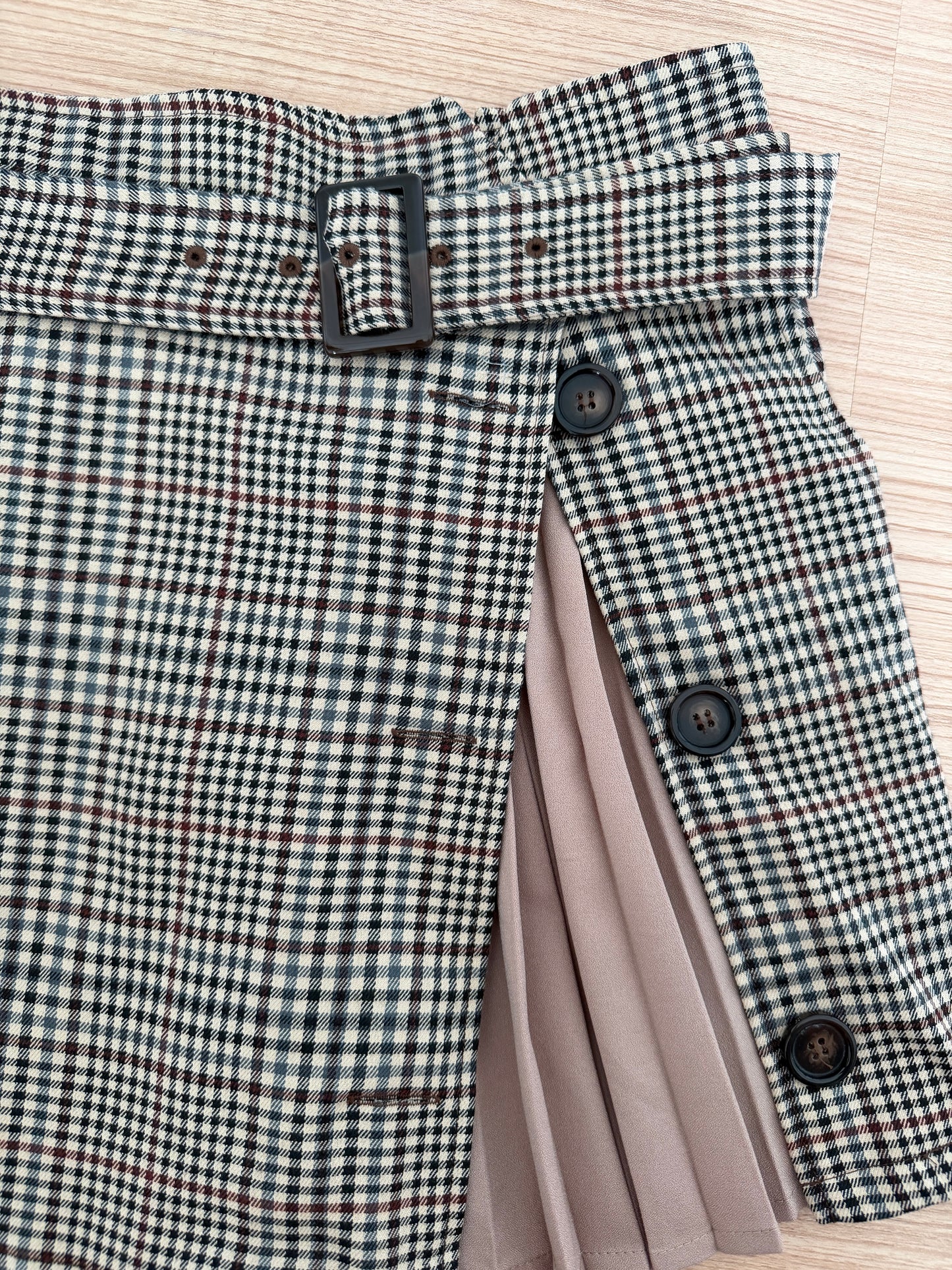 A close-up of a skirt with a black, white, and beige gingham pattern, featuring a matching waist belt with a black buckle, decorative buttons, and a pleated taupe lining visible on the side, laid flat on a light wood background.
