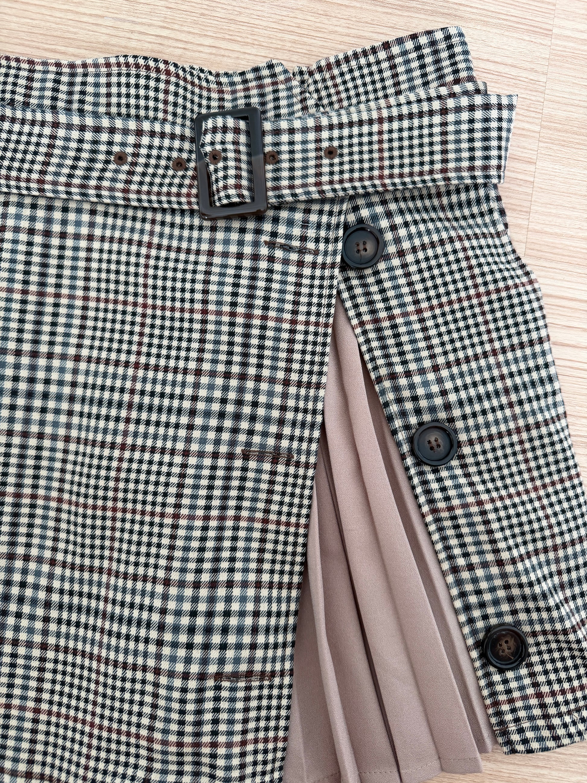 A close-up of a skirt with a black, white, and beige gingham pattern, featuring a matching waist belt with a black buckle, decorative buttons, and a pleated taupe lining visible on the side, laid flat on a light wood background.