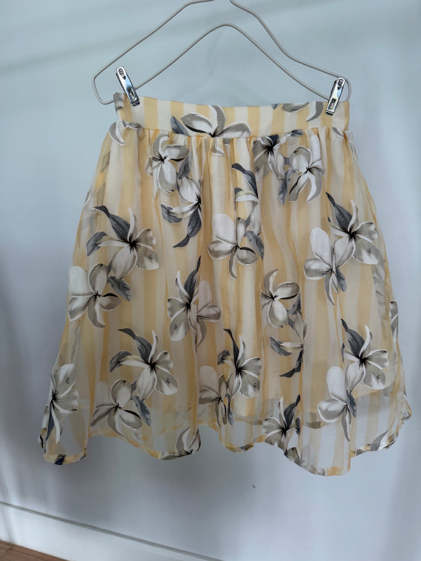 A sheer yellow and white striped mini skirt with a gray and white floral print, hanging from a metal hanger against a white wall.