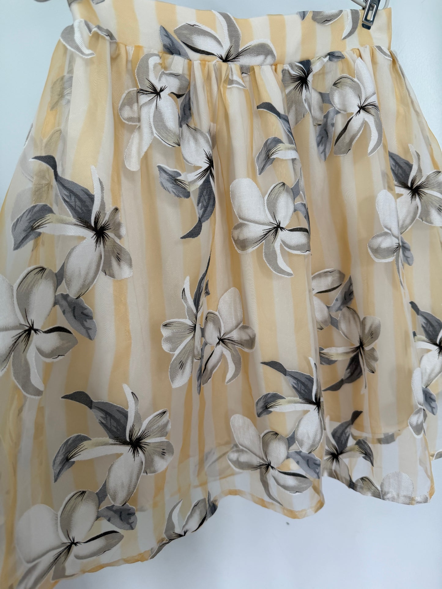 A sheer pleated skirt in a pale yellow and white striped pattern, decorated with a large-scale gray and white floral print, hanging against a plain white background.