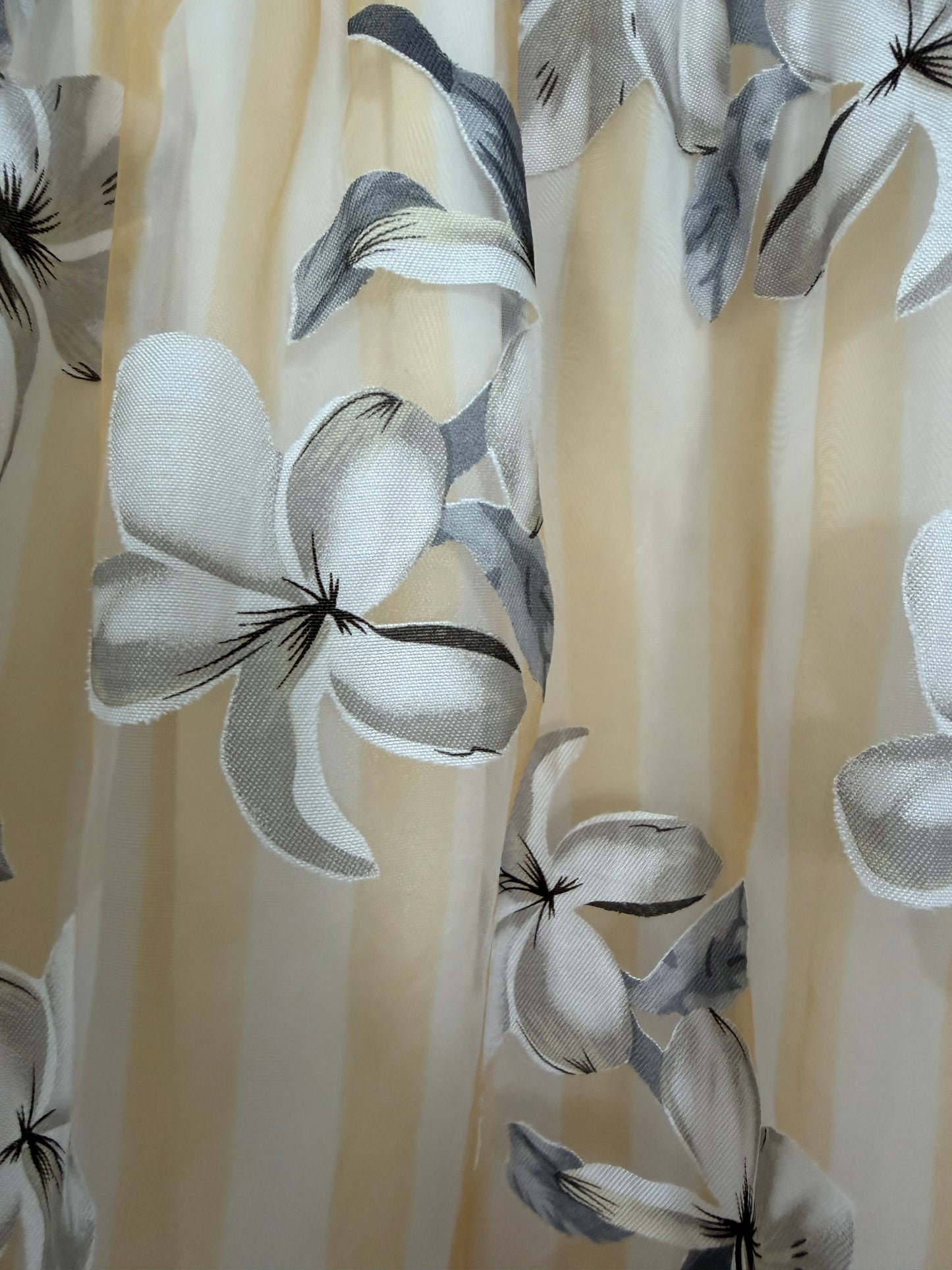 A close-up of a sheer curtain fabric with a vertical striped pattern in cream and beige, decorated with a large-scale floral print of white and gray plumeria flowers with dark accents.