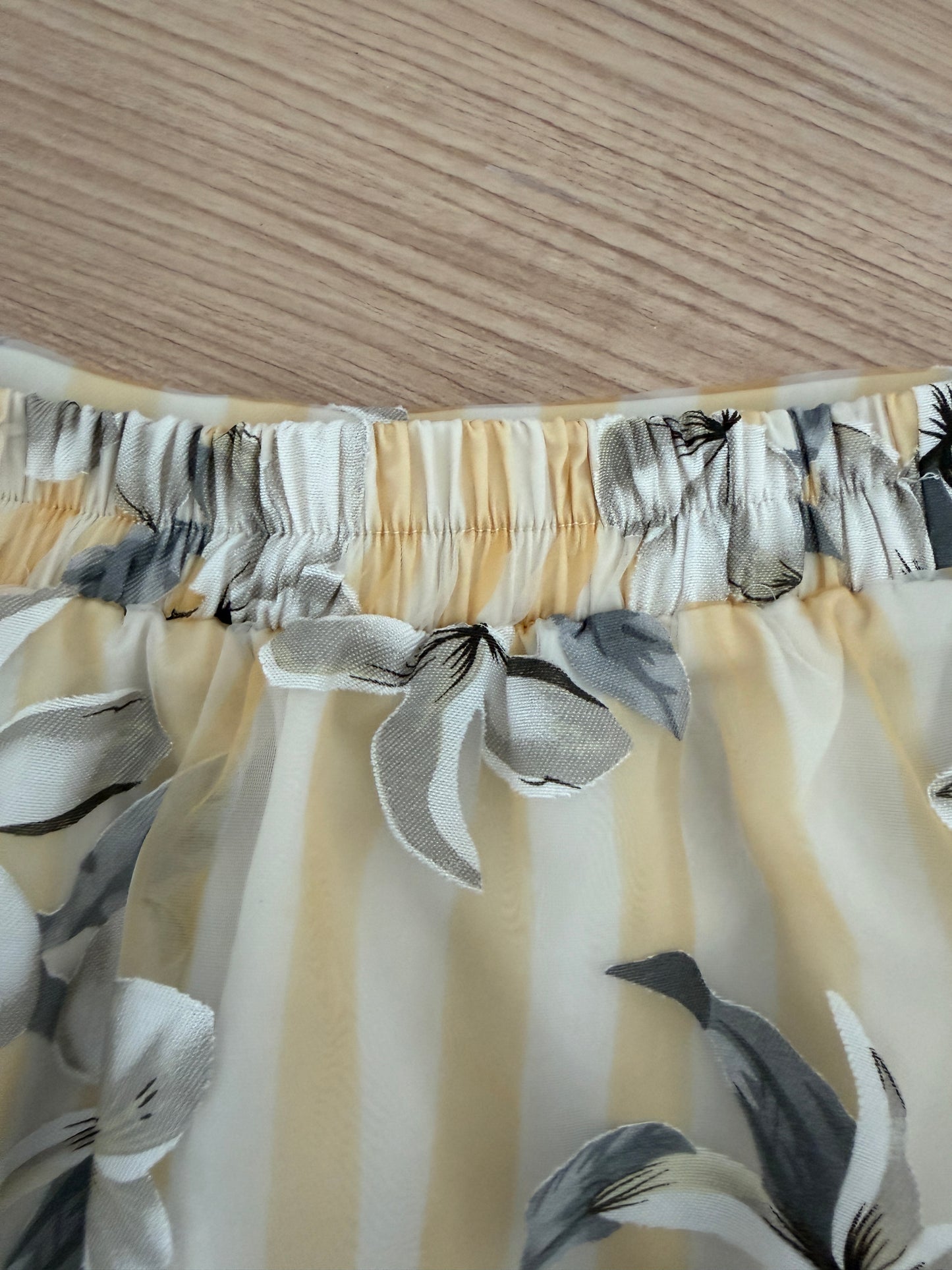 Close-up of a skirt or dress fabric with a gathered elastic waistband, featuring a cream and pale yellow striped base with gray and white floral print and bow details.