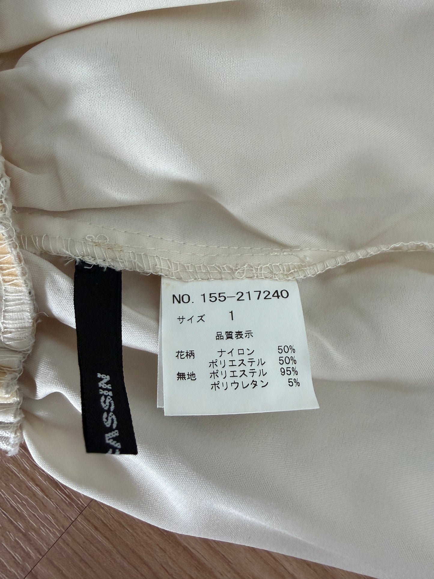 Close-up of a garment's interior label area showing a white size tag and a black NISSIN brand tag attached to cream-colored satin fabric.