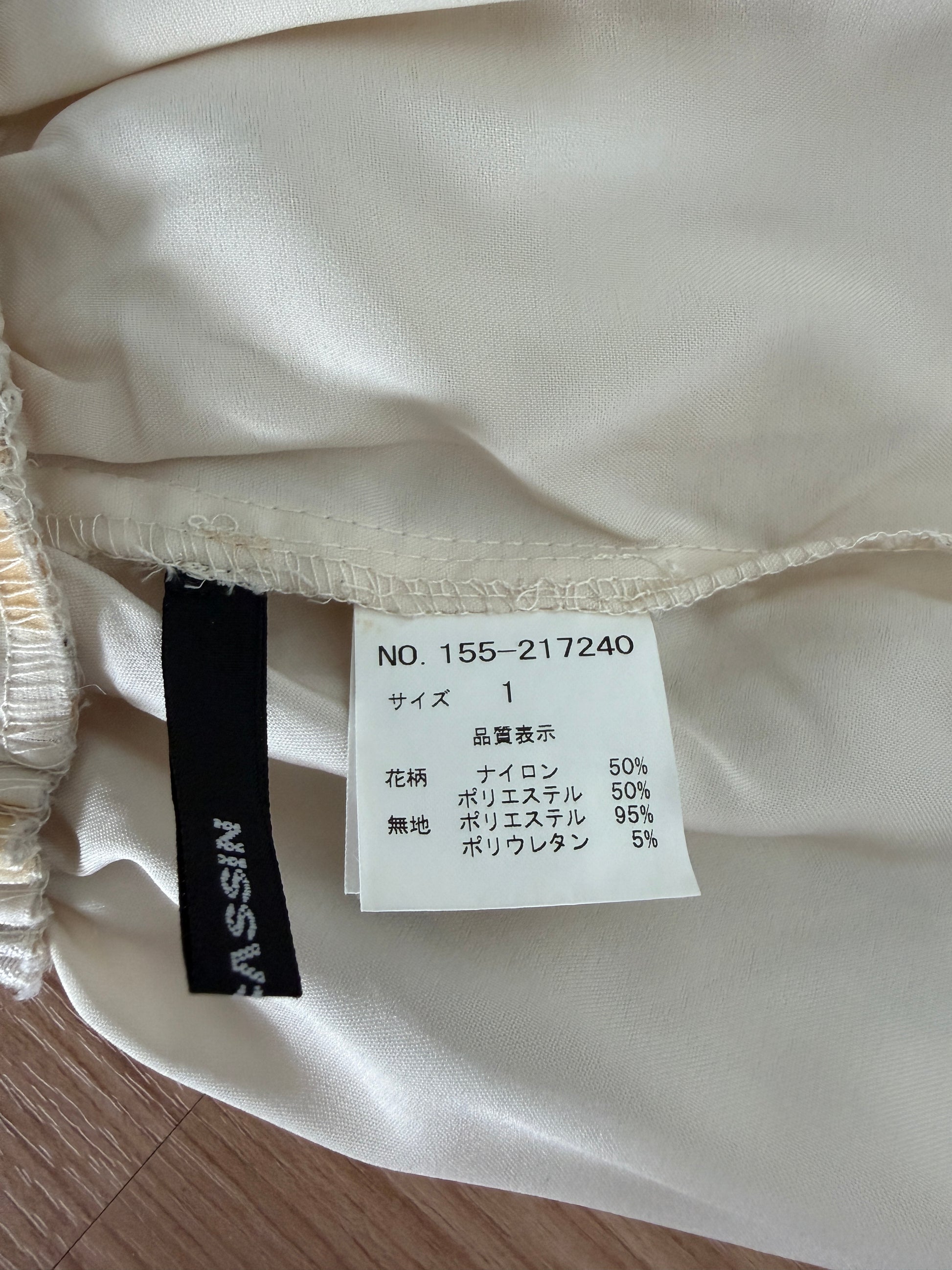 Close-up of a garment's interior label area showing a white size tag and a black NISSIN brand tag attached to cream-colored satin fabric.