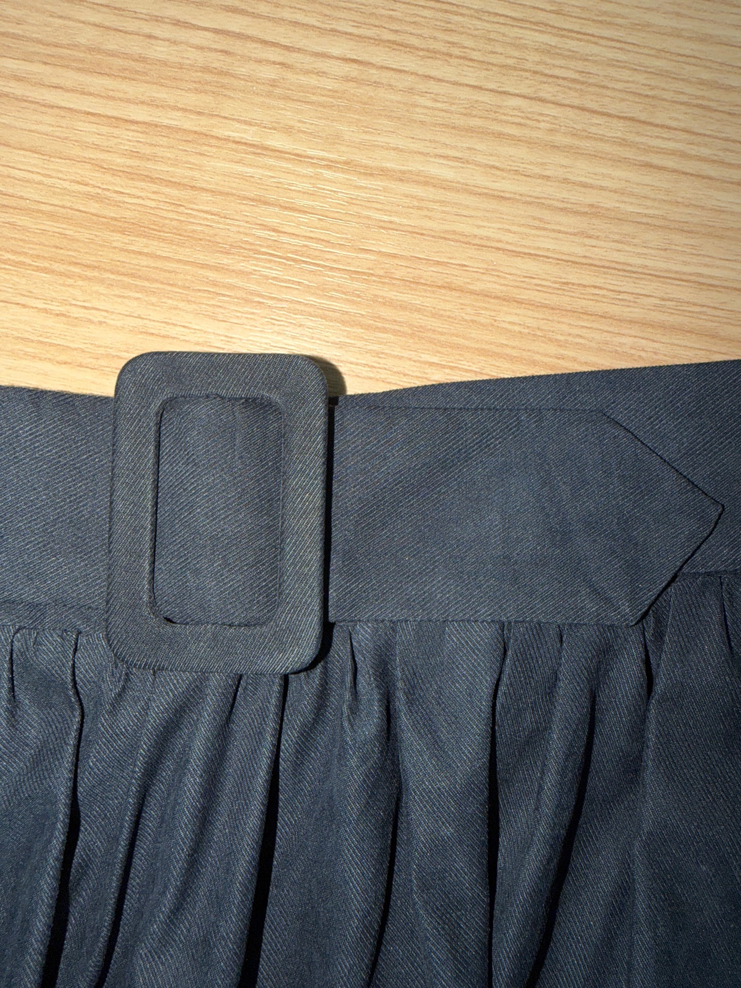 Close-up of a dark gray pleated skirt with a wide matching belt and a square buckle, laid flat on a light wood surface.