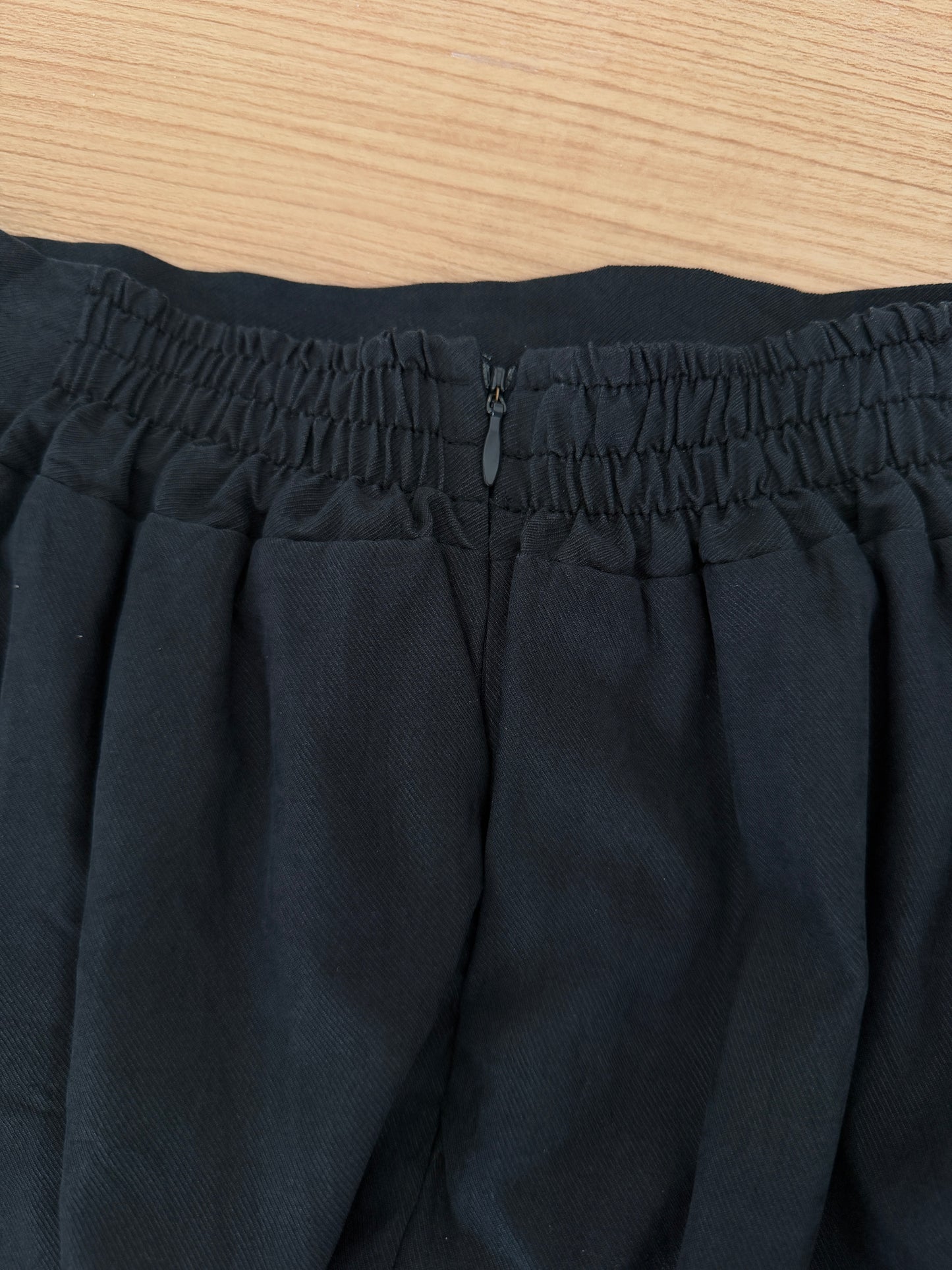 Close-up of black pleated fabric with an elasticized waistband and a small metal zipper pull, laid flat on a light wood surface.