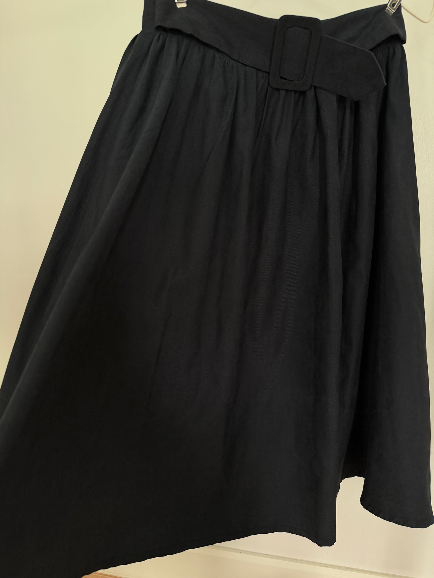 A black high-waisted A-line skirt with a wide matching belt and a rectangular buckle detail, shown hanging against a plain light beige wall.
