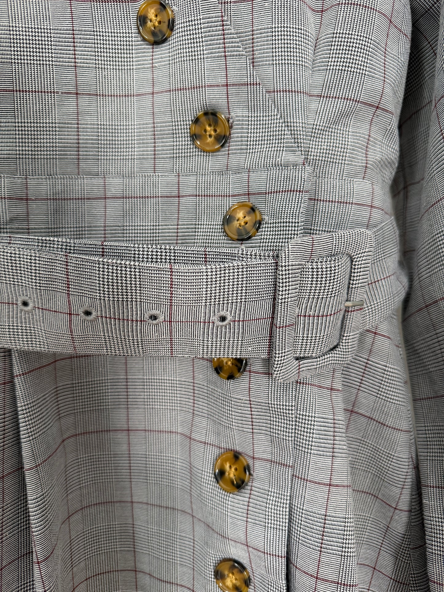 Close-up of a belted jacket or coat with a fine black and white houndstooth pattern accented by thin red grid lines and large tortoiseshell buttons.
