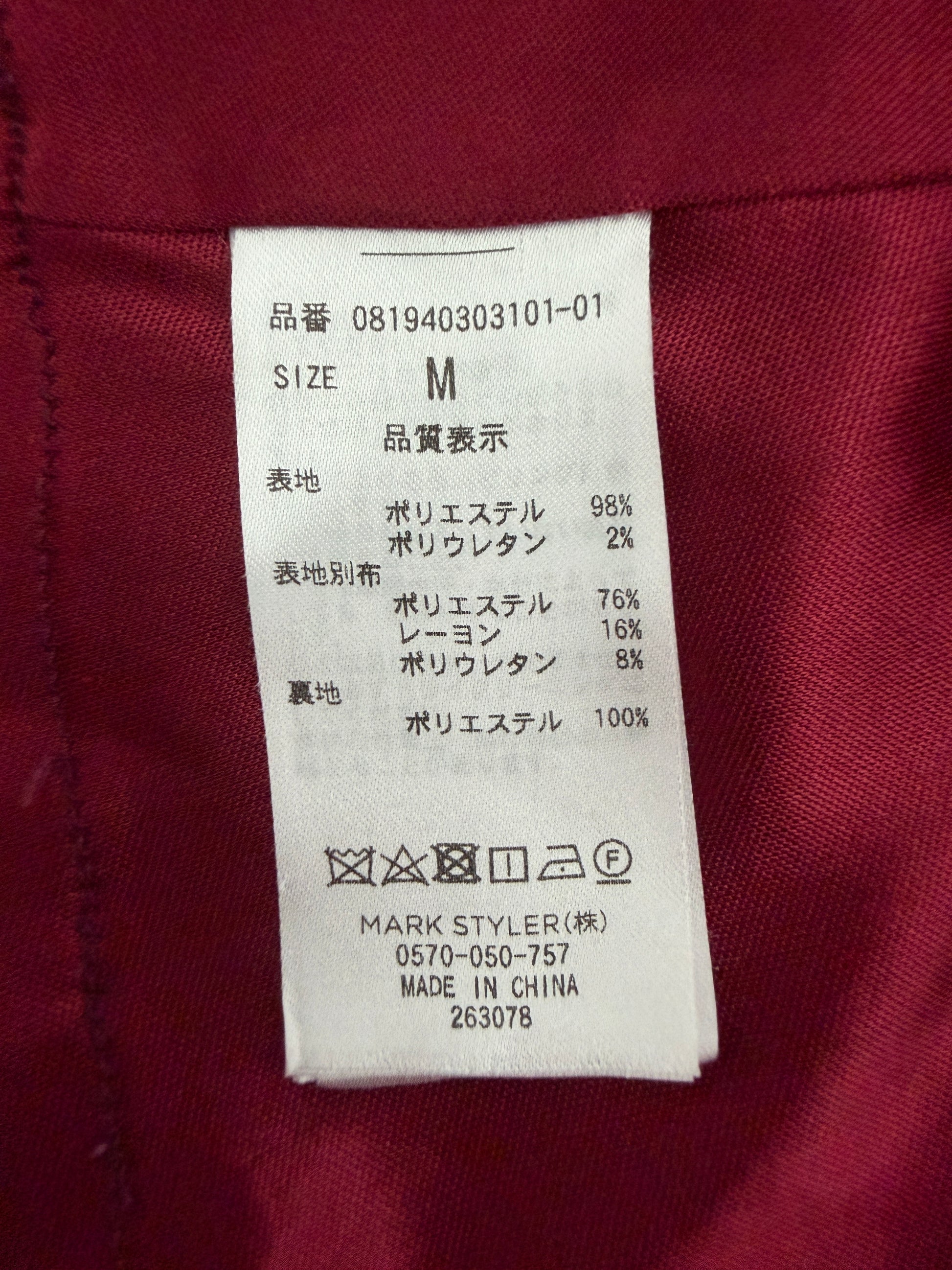 A close-up of a garment's interior label sewn onto a dark burgundy fabric. The white rectangular label shows size M and material composition details in Japanese, including 98% polyester, 2% polyurethane, 76% polyester, 18% lycra, and 8% elastic, with the brand name MARK STYLER visible at the bottom.