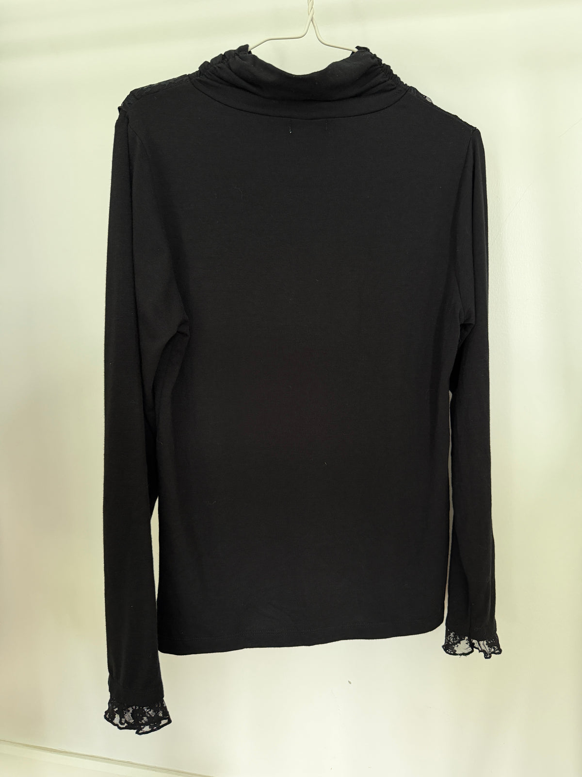 A black long-sleeve turtleneck top with lace trim at the cuffs, hanging on a white hanger against a plain off-white background.