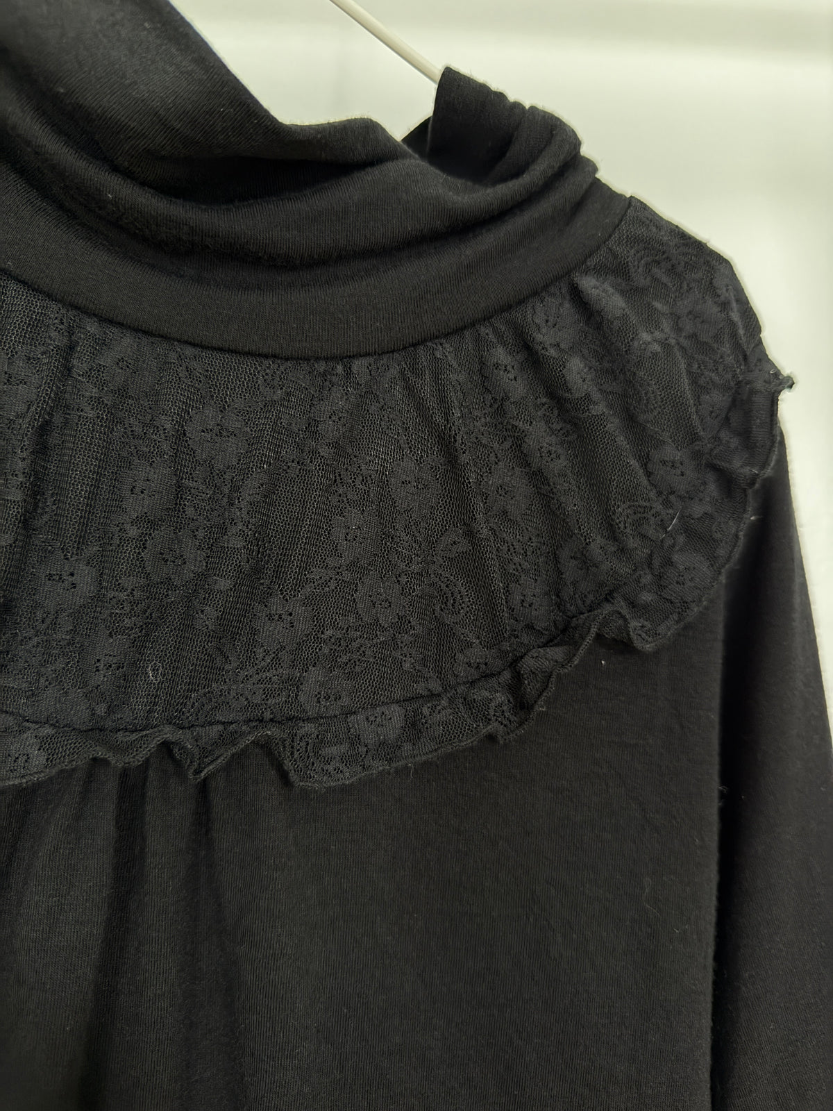 Close-up of a black long-sleeve top with a wide ruffled lace collar and a folded cowl neckline, shown hanging against a light neutral background.
