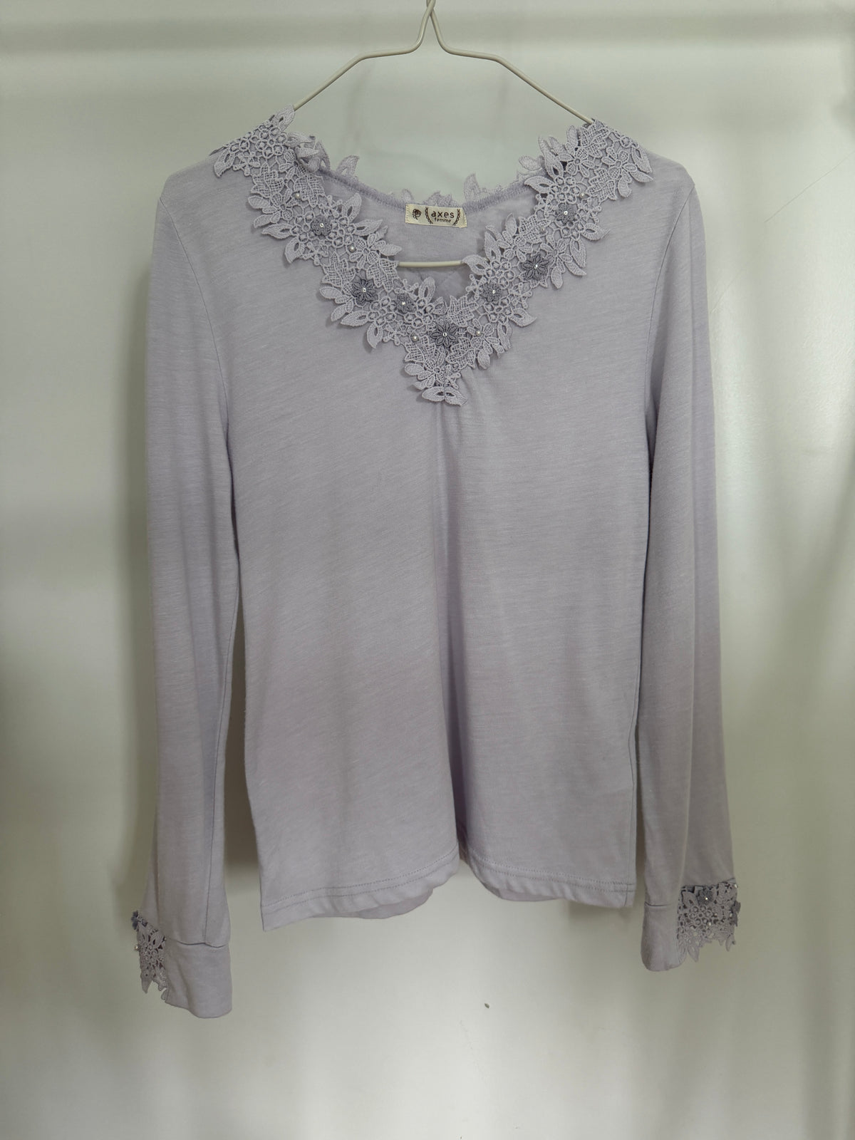 A light lavender long-sleeve top with a floral lace V-neckline and matching lace cuffs, hanging on a metal hanger against a plain light background.