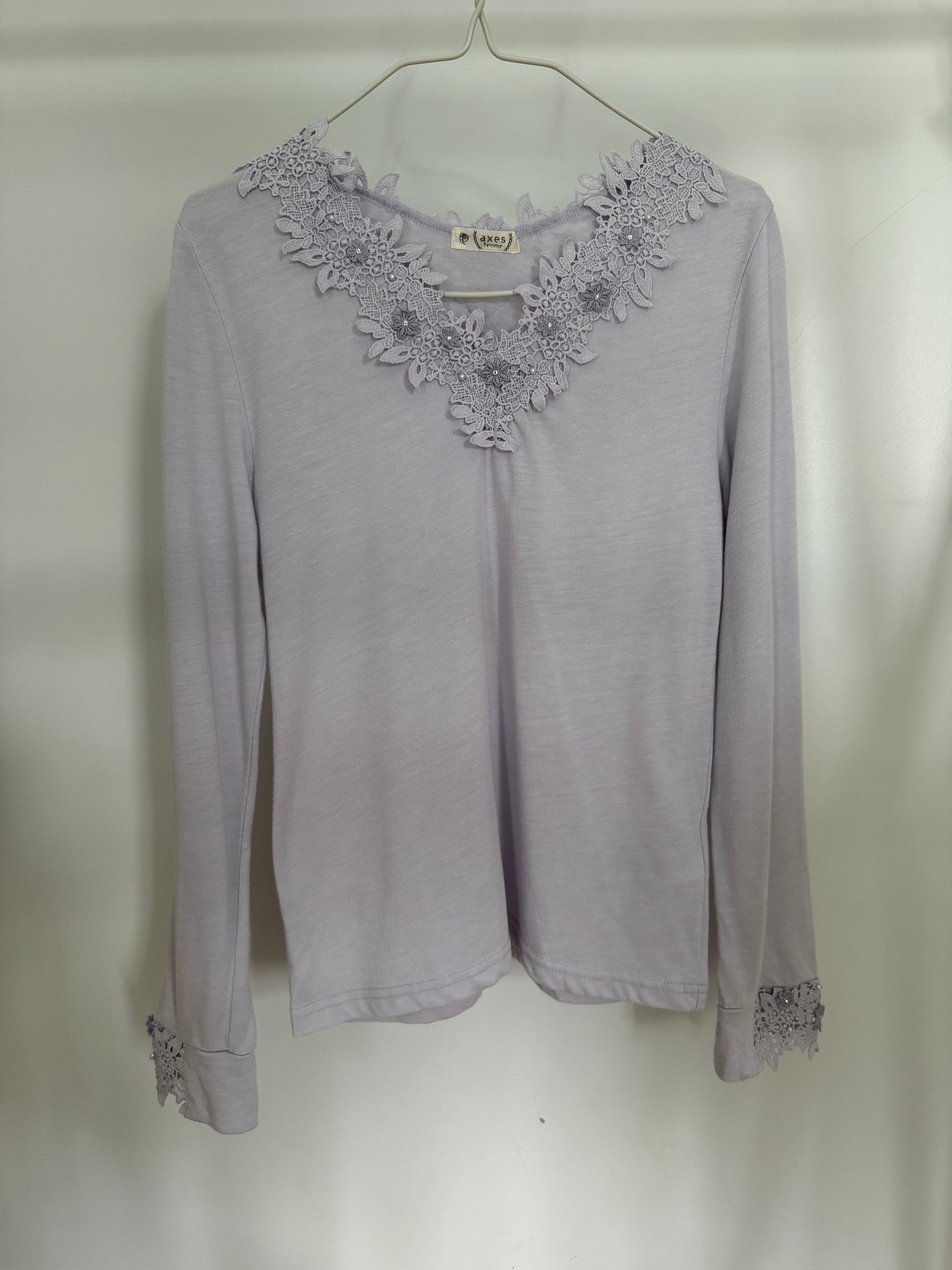 A light lavender long-sleeve top with a floral lace V-neckline and matching lace cuffs, hanging on a metal hanger against a plain light background.