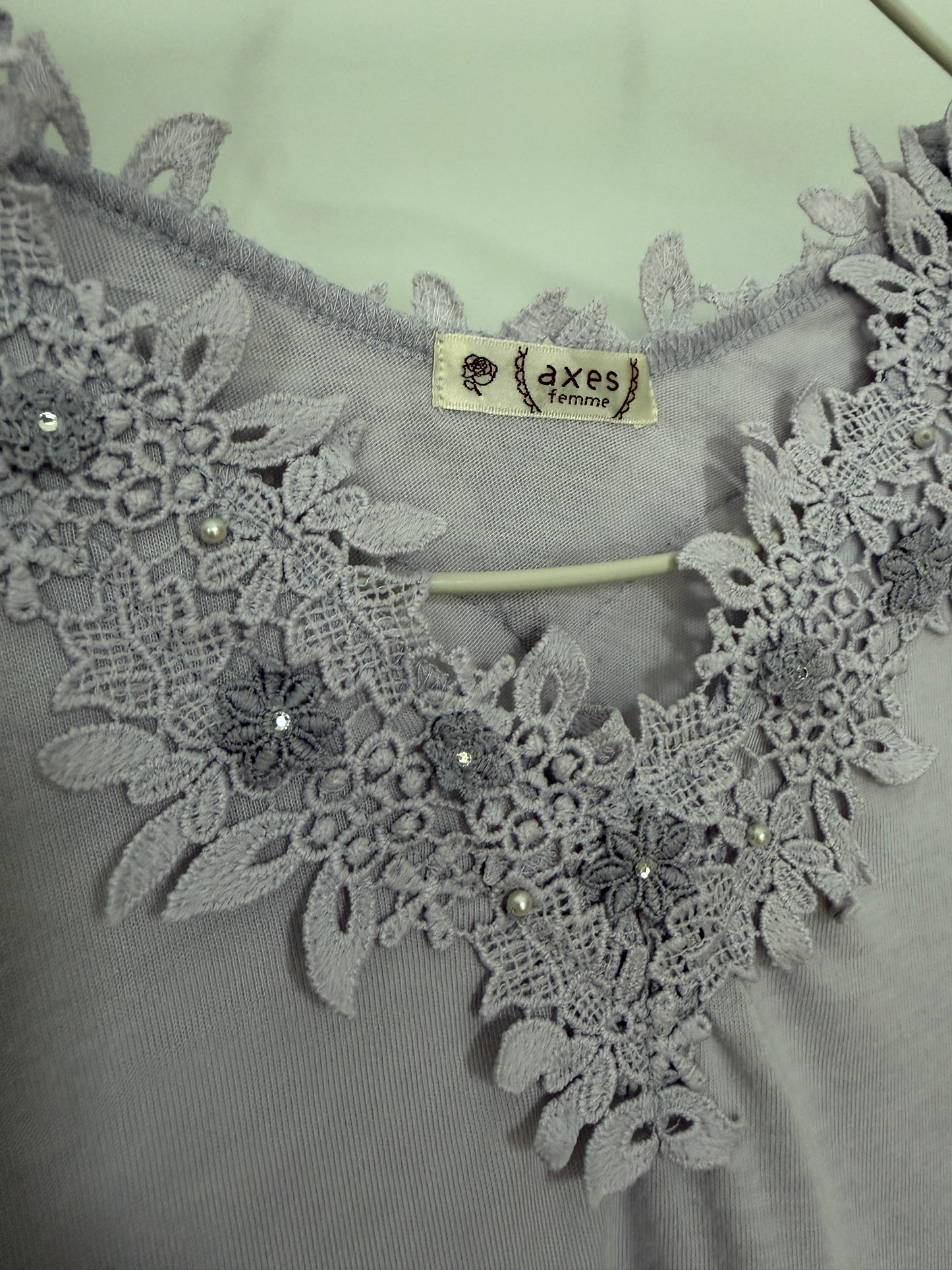 Close-up of a light gray blouse featuring an intricate floral lace neckline with small pearl embellishments and a visible 'Akses' brand label.