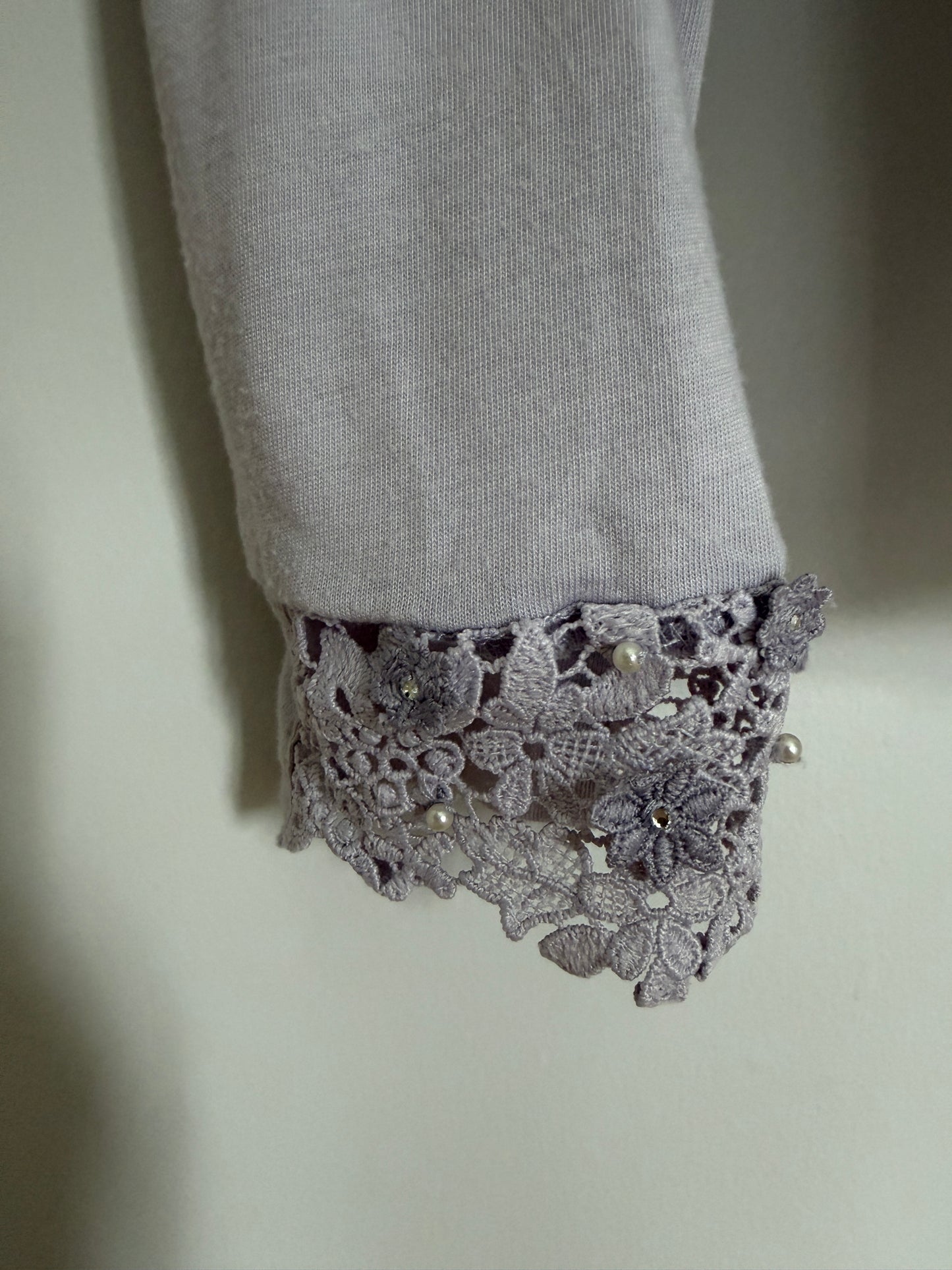 Close-up of a light gray fabric sleeve with a decorative lace cuff featuring small pearl-like beads and floral lace detailing.
