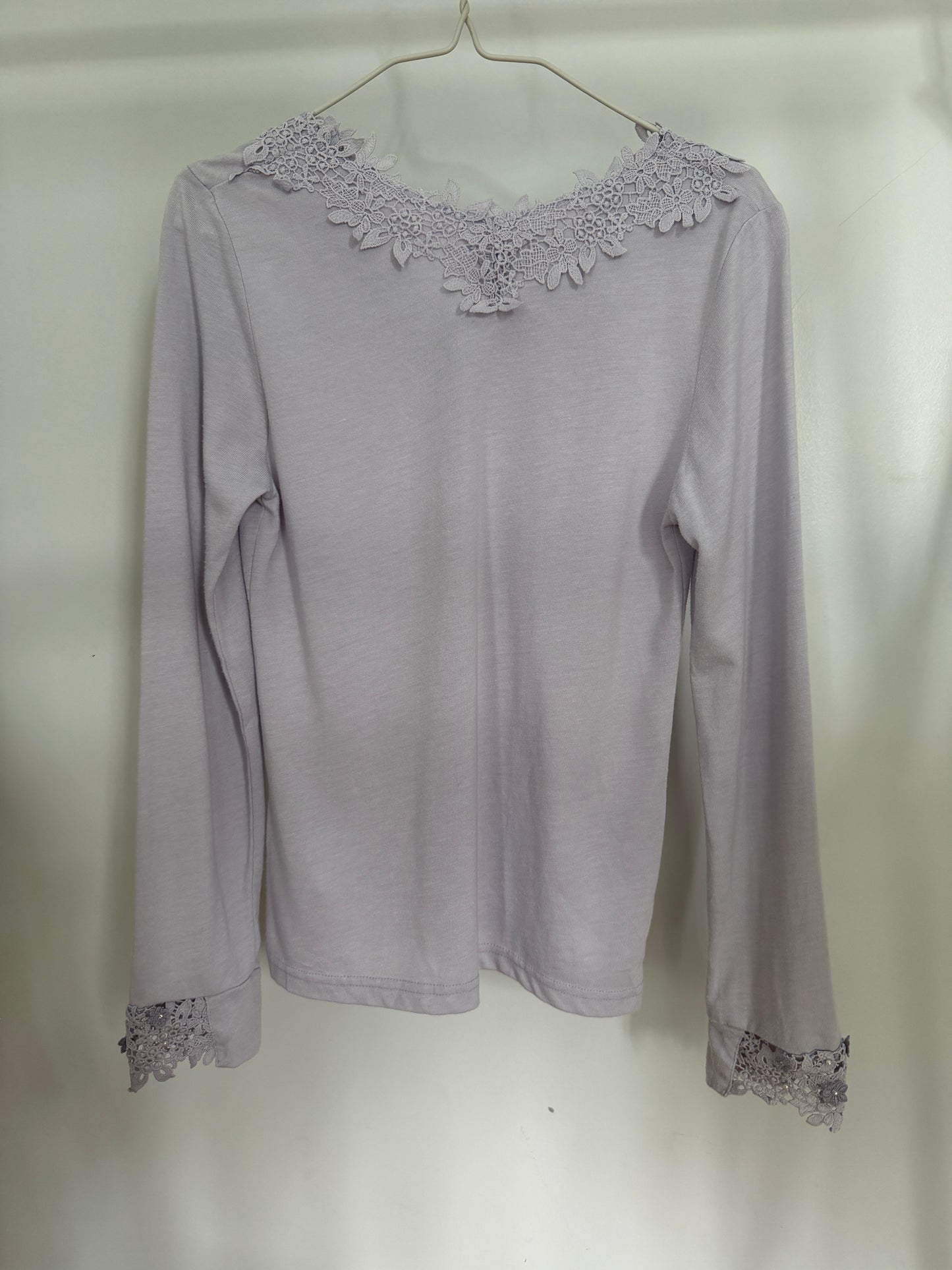 A light lavender-gray long-sleeve top with floral lace trim at the round neckline and cuffs, hanging on a metal hanger against a plain light background.
