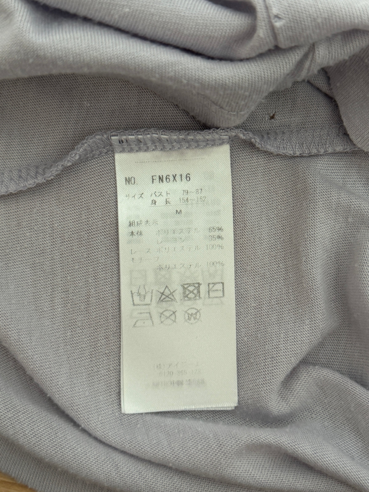 Close-up of a light gray garment's interior label sewn onto the inside neck area, showing product code FN6X16 and care symbols on a white paper tag.