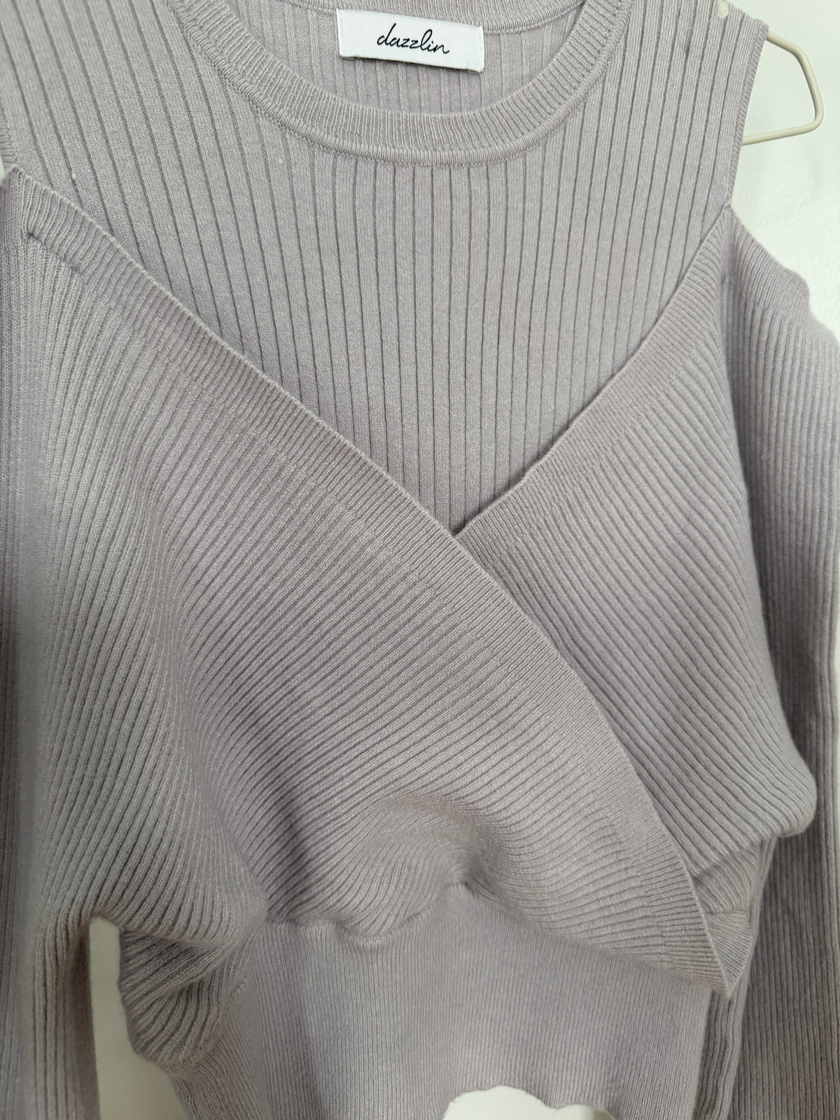 A close-up of a light gray ribbed knit top with a crossover wrap-style front and long sleeves, hanging on a hanger. The brand name 'dazzler' is visible on the inner neck label.