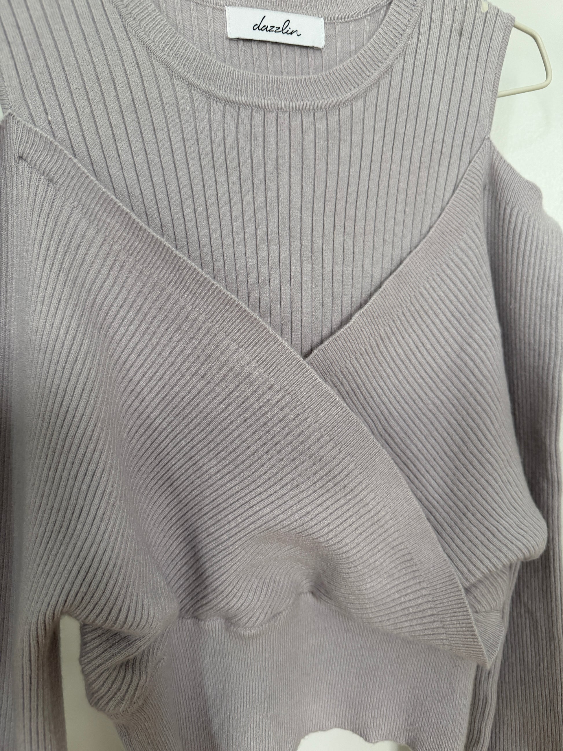 A close-up of a light gray ribbed knit top with a crossover wrap-style front and long sleeves, hanging on a hanger. The brand name 'dazzler' is visible on the inner neck label.