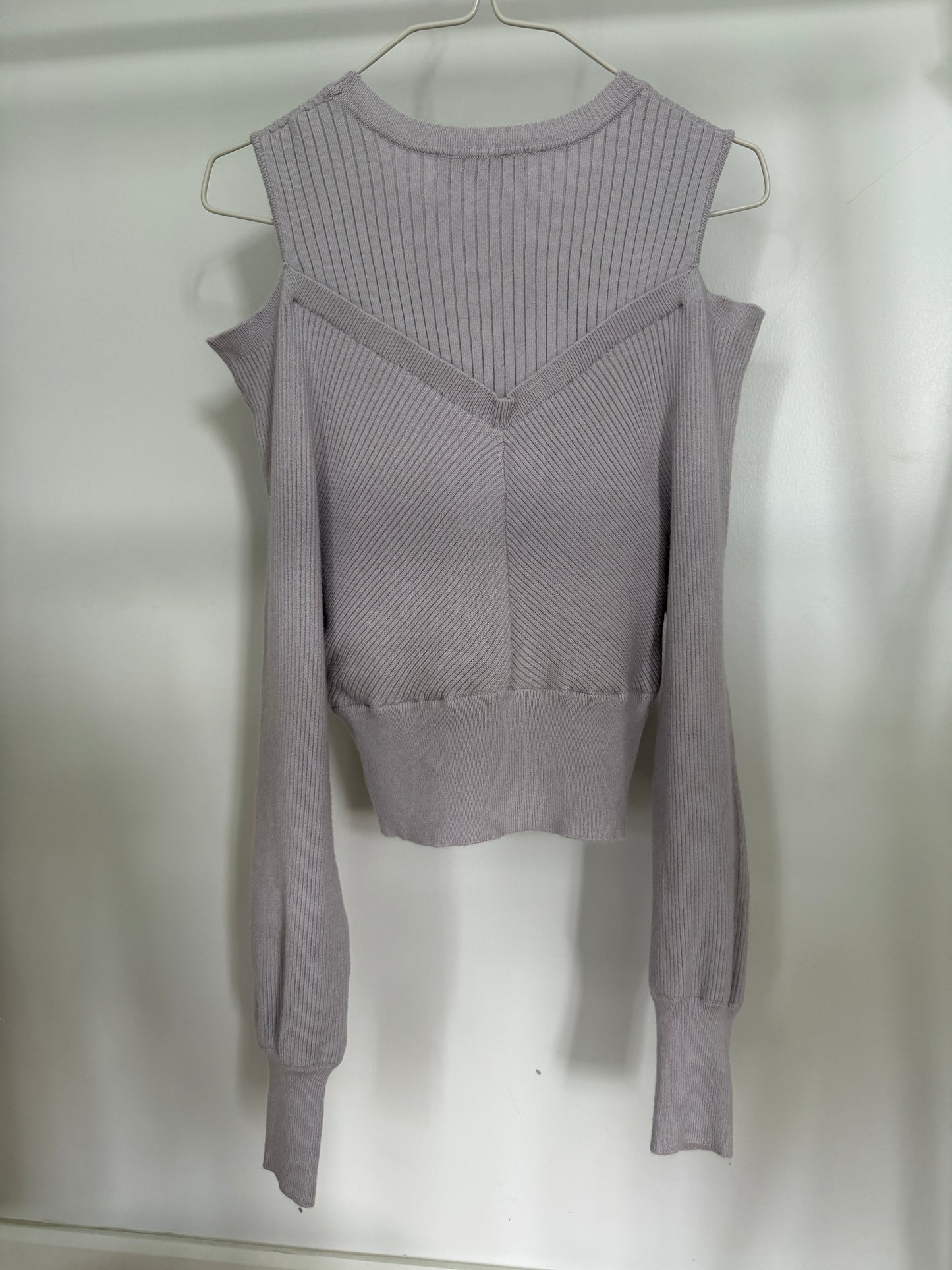 A light gray ribbed knit long-sleeve top with a V-neckline and shoulder cutouts, hanging on a metal hanger against a plain white wall.