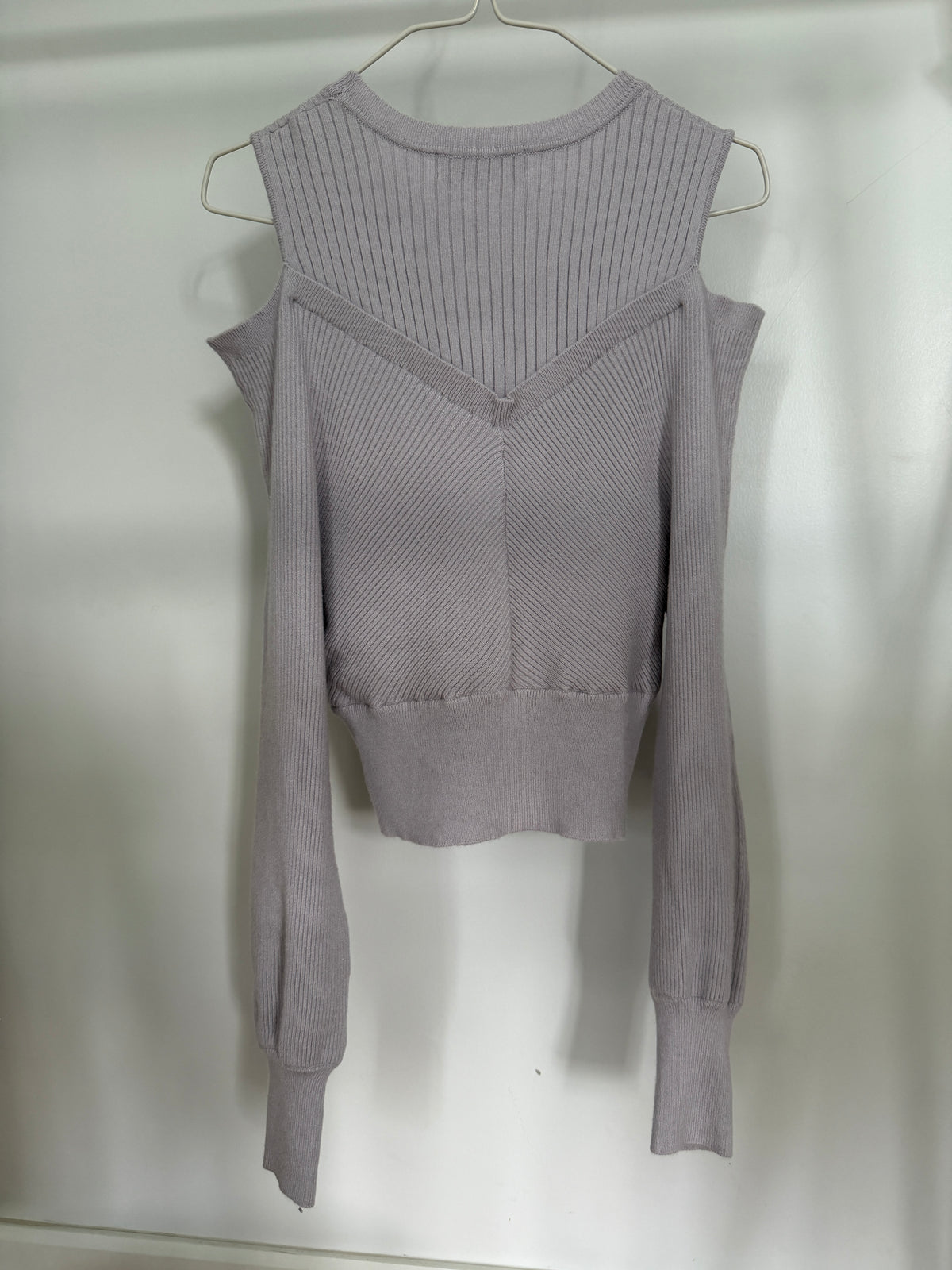 A light gray ribbed knit long-sleeve top with a V-neckline and shoulder cutouts, hanging on a metal hanger against a plain white wall.