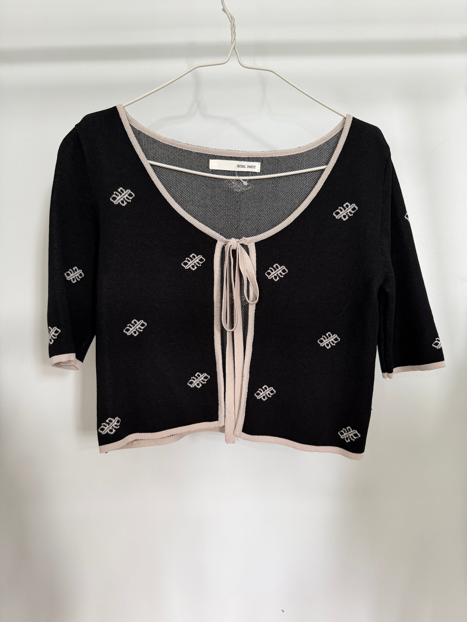 A black knit cardigan top with beige trim and a front tie closure, hanging on a white wire hanger against a white wall. The top features small white geometric patterns throughout.