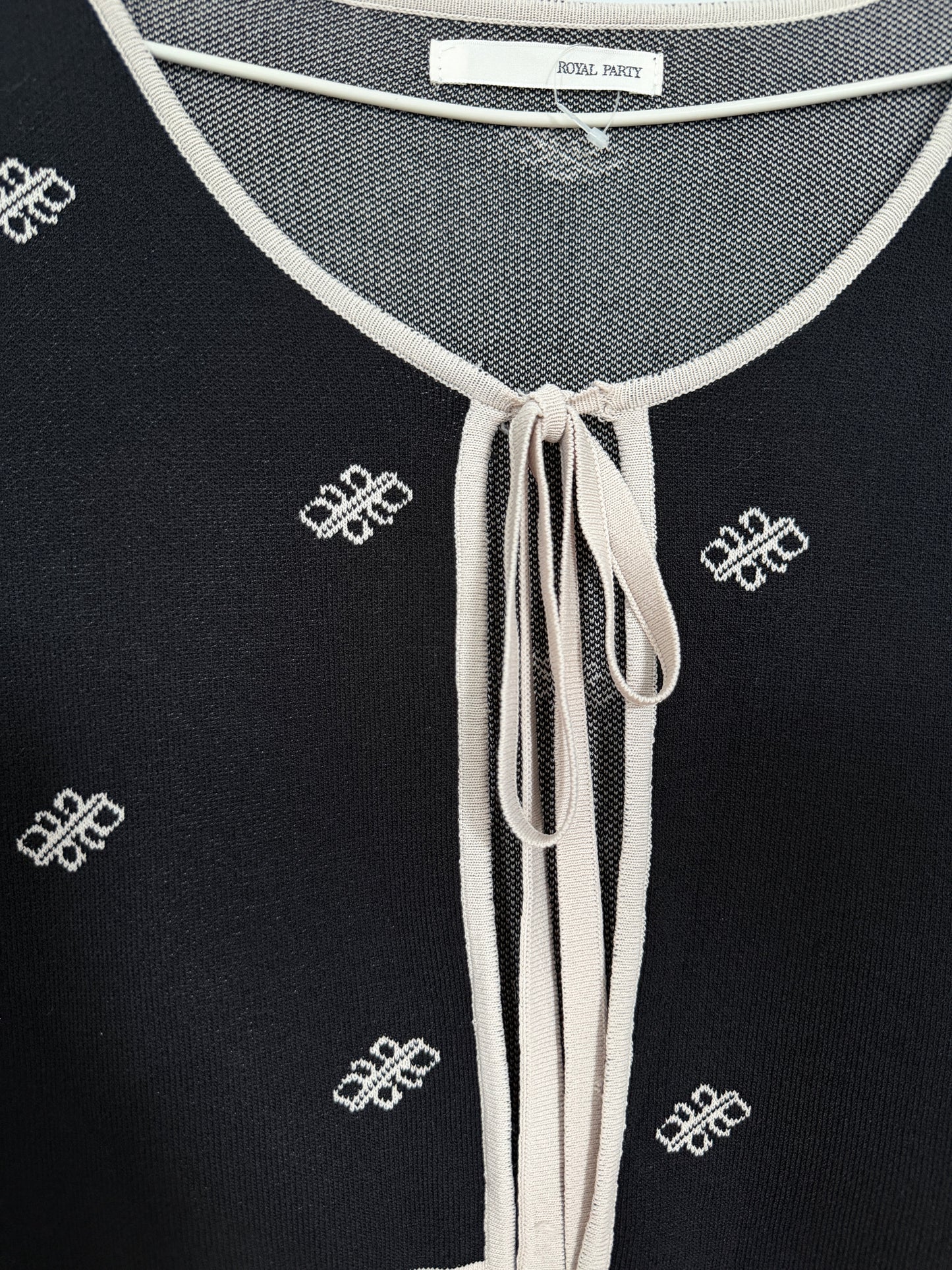 Close-up of a black knit top with a cream tie-neck trim and small white embroidered geometric motifs, showing a neck label that reads 'ROYAL PARTY'.