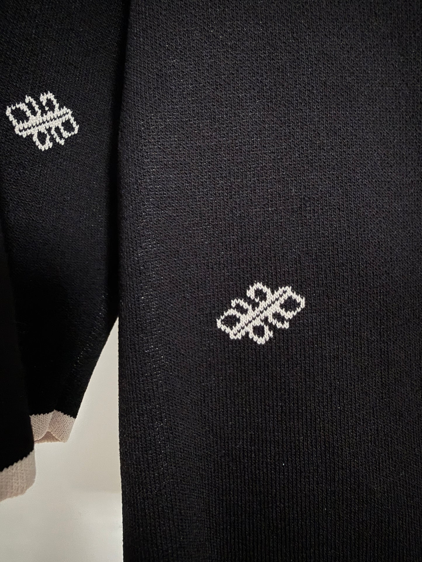 Close-up of a black knit garment, likely a sweater or cardigan, featuring a small white geometric embroidered motif on the chest and sleeve.
