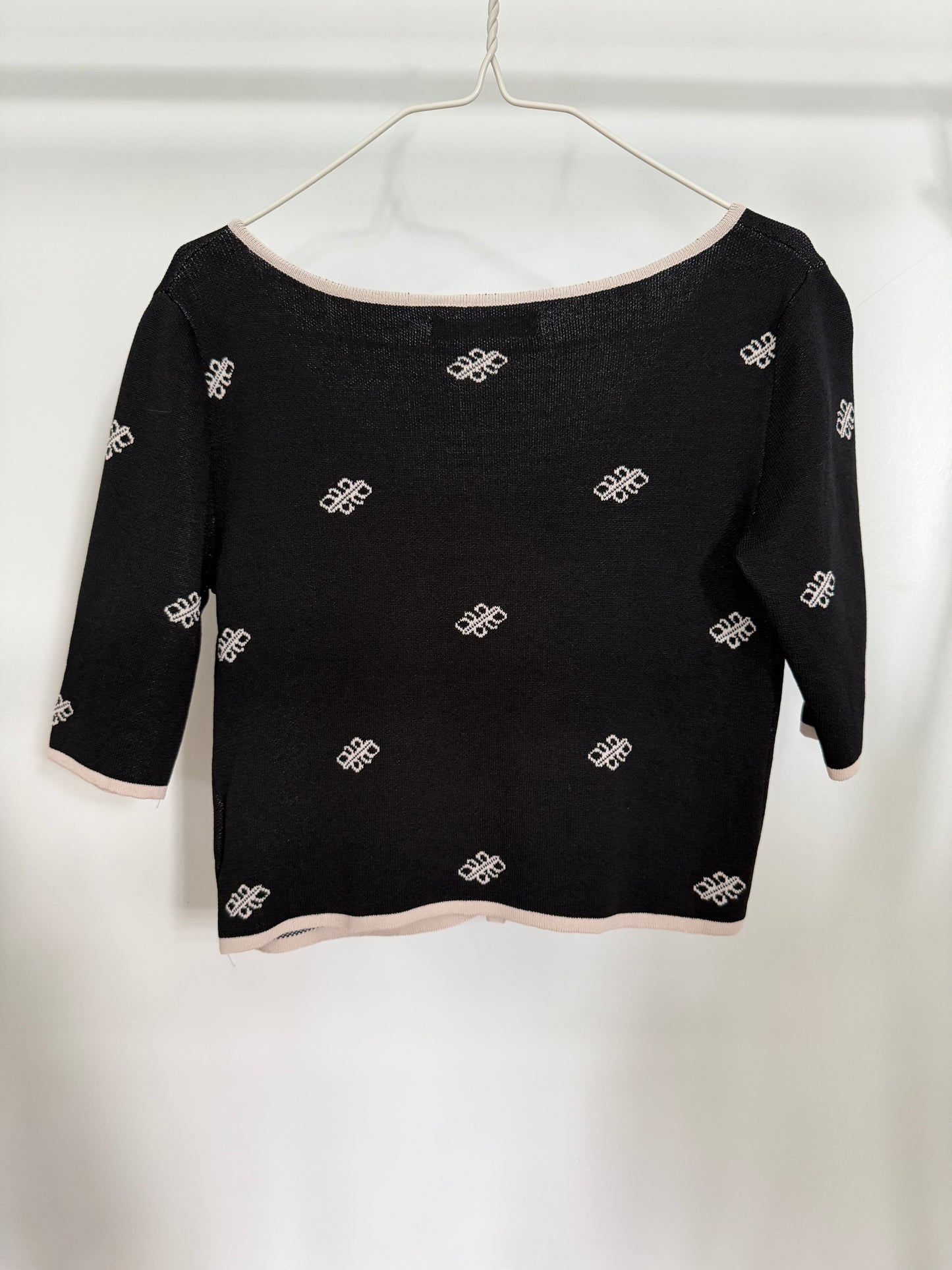 A black knit off-the-shoulder top with three-quarter sleeves, featuring a repeating white embroidered monogram pattern and light pink trim along the neckline, cuffs, and hem, hanging on a white hanger against a white background.