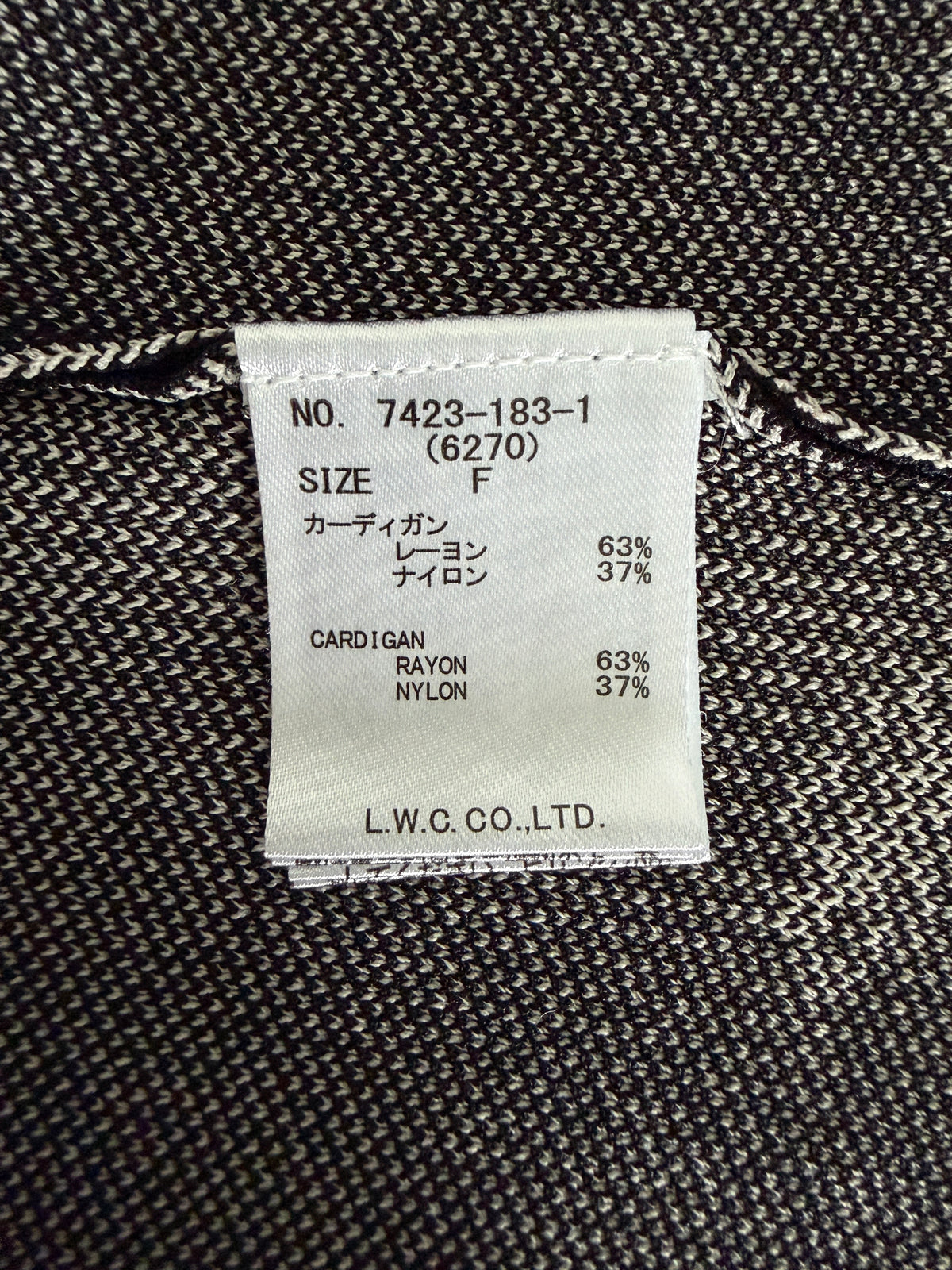A close-up of a garment's interior label sewn onto dark gray and black textured knit fabric. The white paper label shows text including 'NO.