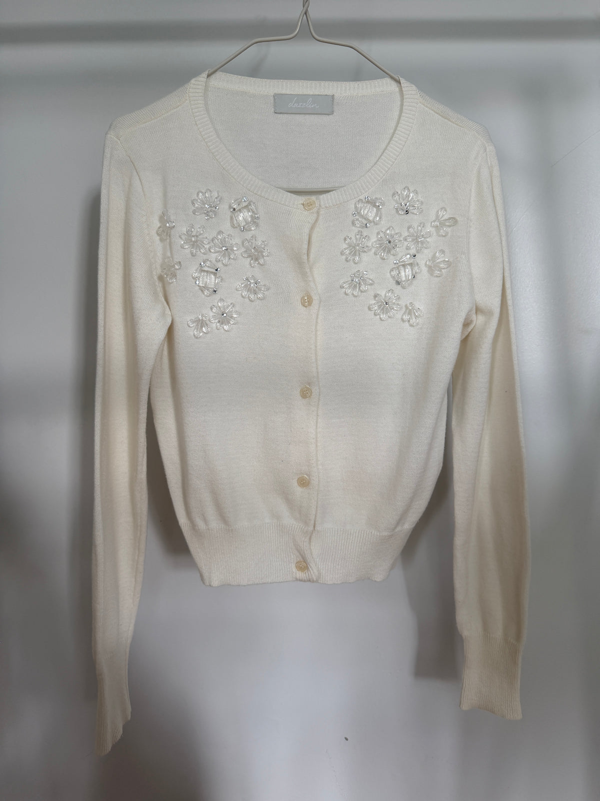 An off-white long-sleeve cardigan sweater with floral embellishments on the chest, hanging on a metal hanger against a light gray background.