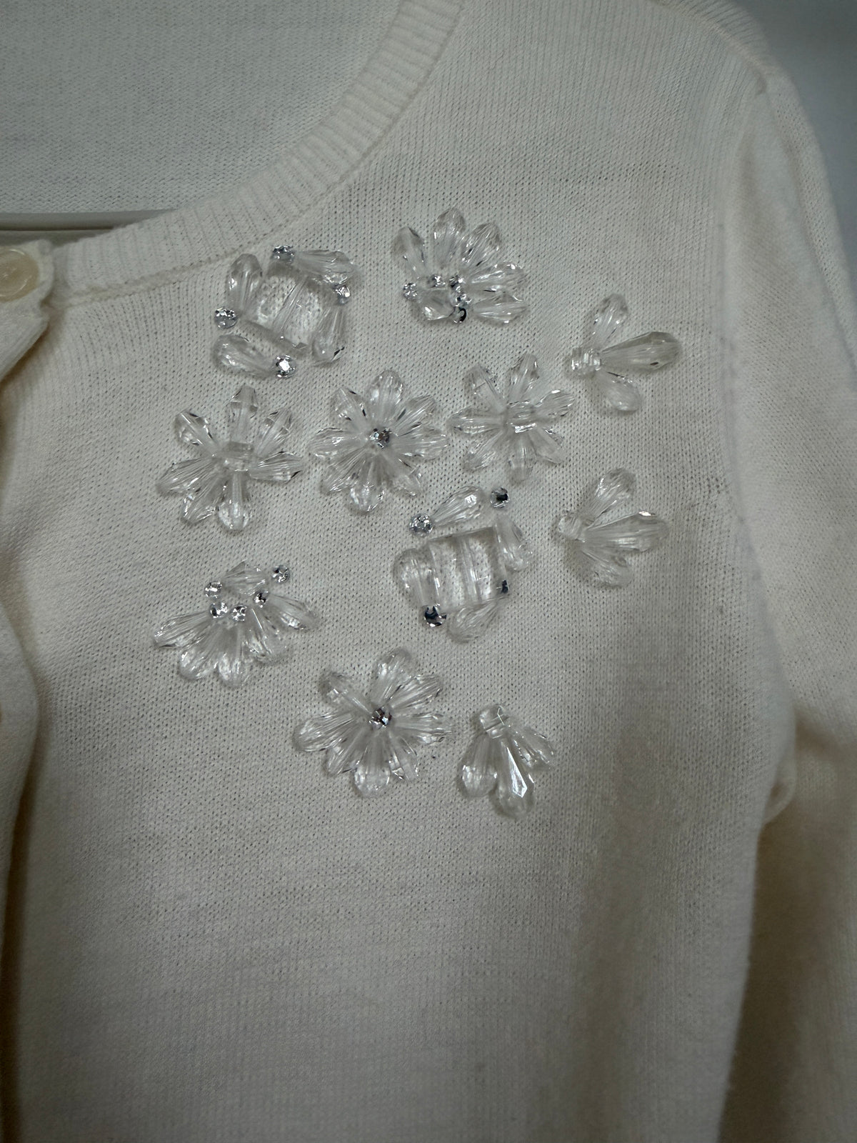 Close-up of a cream-colored knit cardigan featuring decorative clear plastic flower appliqués embellished with small rhinestones on the chest.