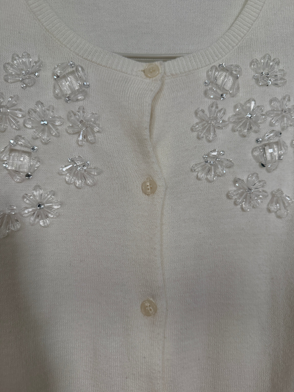 Close-up of a cream-colored knit cardigan featuring clear rhinestone floral embellishments on the chest and buttons down the front.