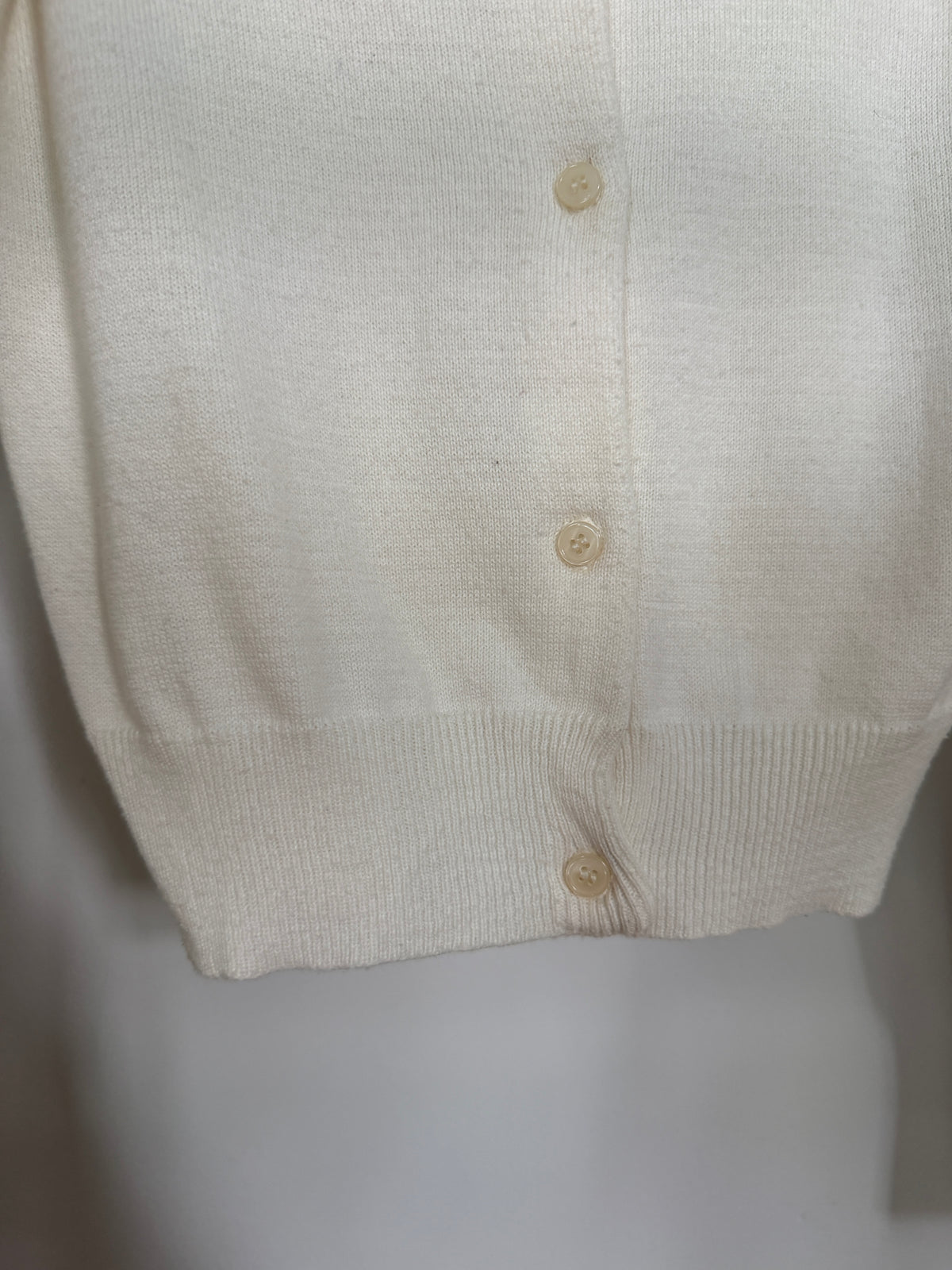 Close-up of a cream-colored knit cardigan sweater showing the ribbed cuffs and hem, with three matching buttons on the front.