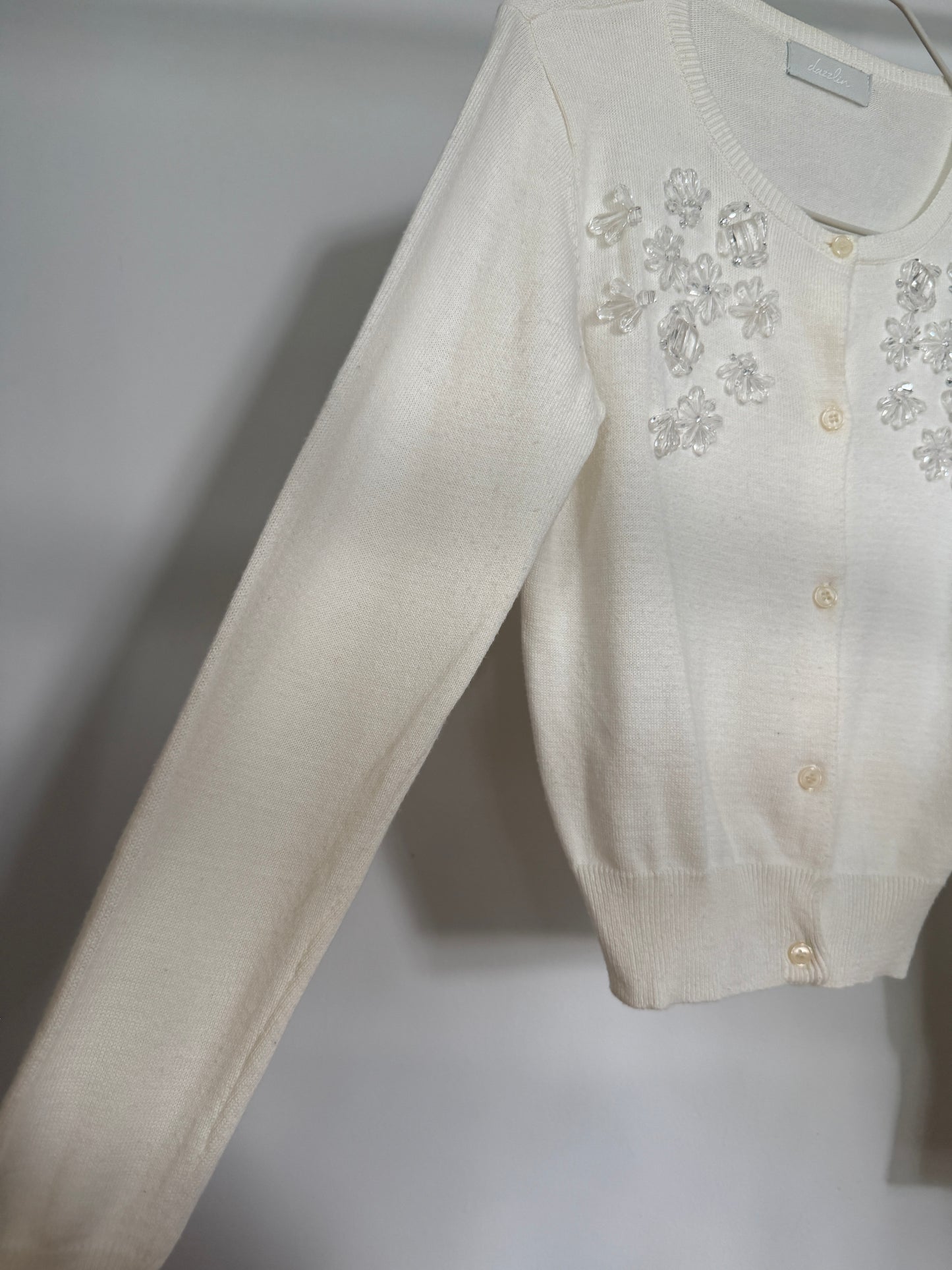 An off-white knit cardigan sweater with floral embellishments and buttons on the chest, shown hanging against a neutral gray background.